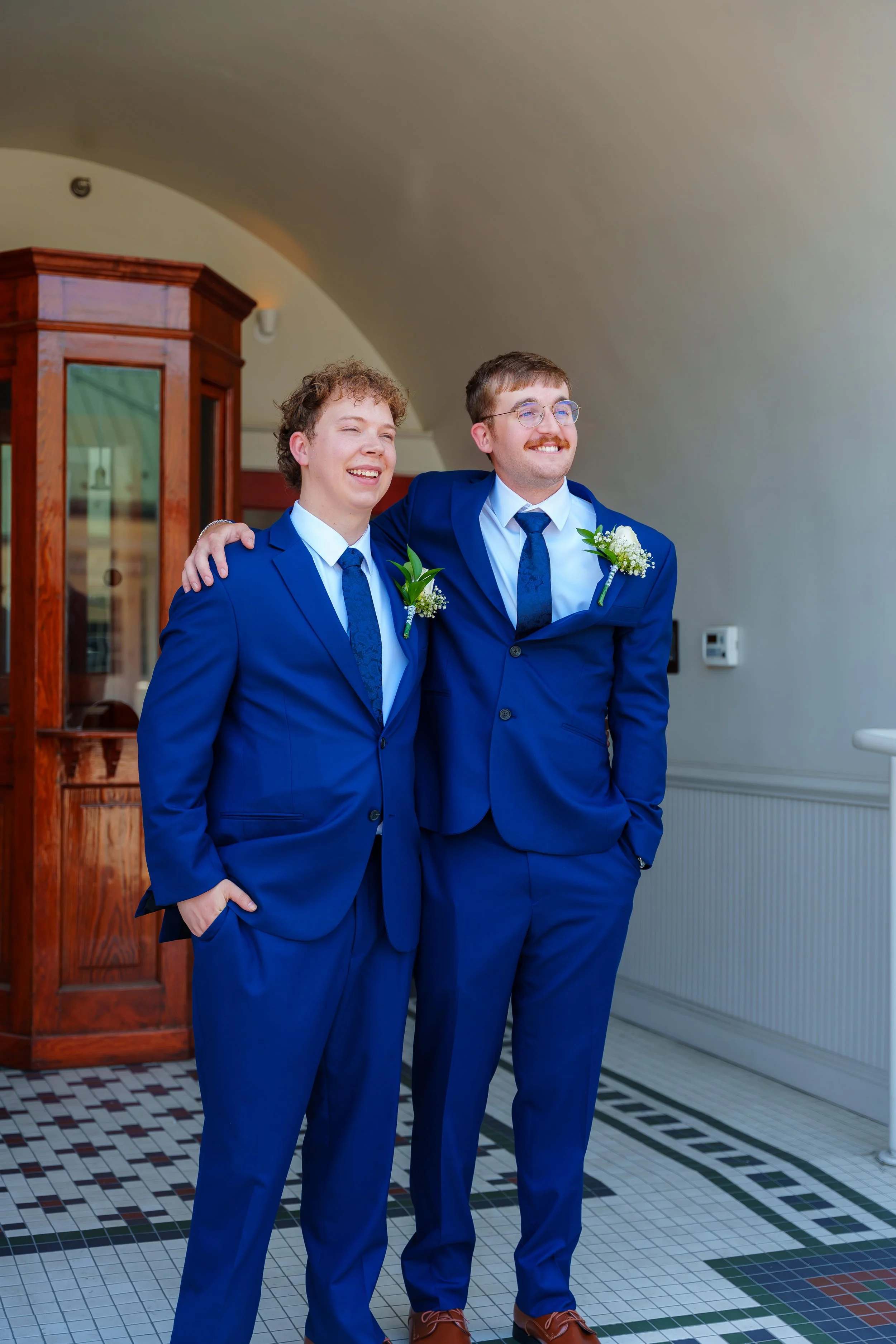 Two young men in matching royal blue suits, white shirts, and blue ties standing close together indoor, smiling, and one has his arm around the other's shoulder. They are both wearing boutonnières with white flowers and greenery. The background inclu