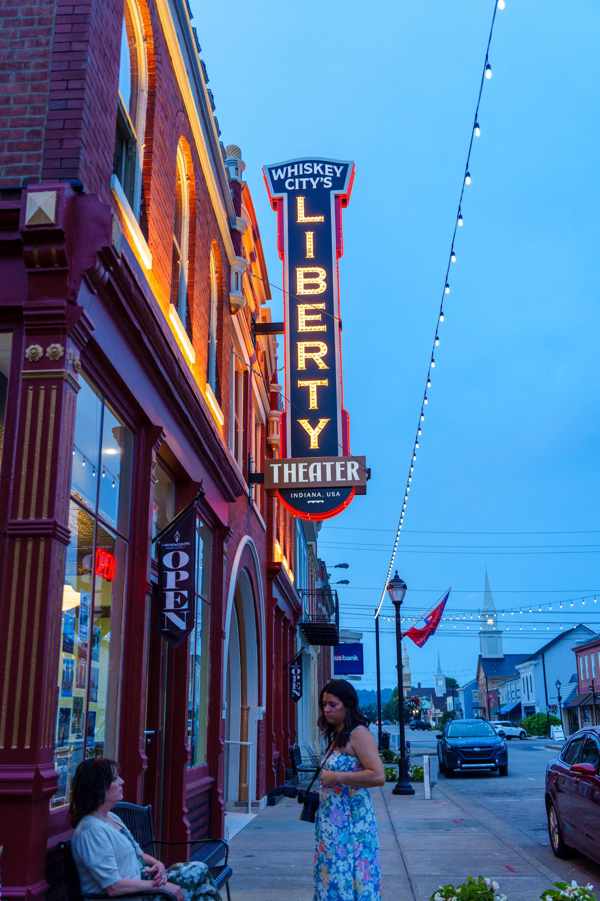 People sitting and standing outside a colorful building with neon sign reading 'Whiskey City' and 'Library' in the evening, with string lights overhead and cars parked on the street.
