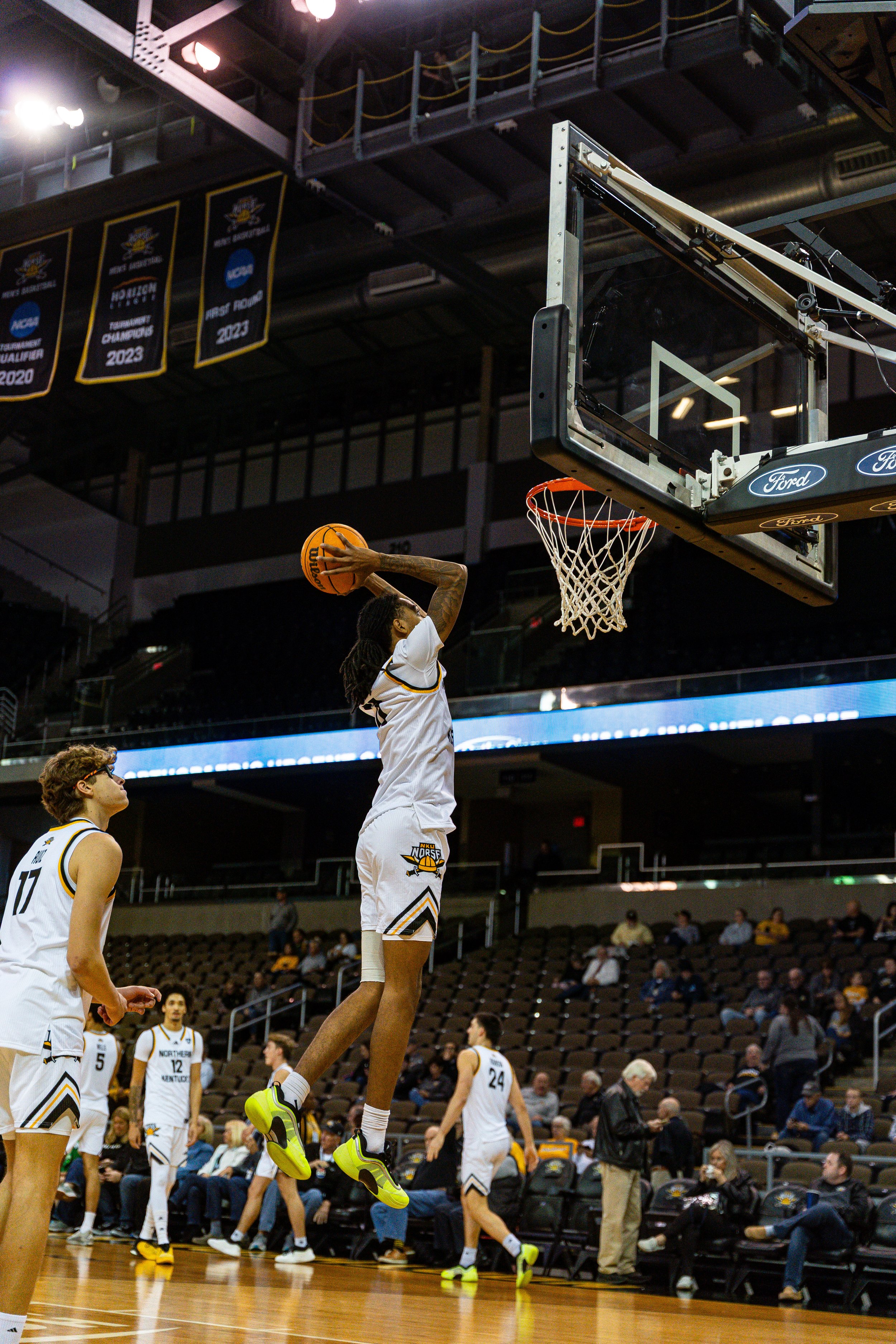 A basketball player in white uniform jumping to shoot the ball towards the basket during a game, with teammates and spectators in the background.