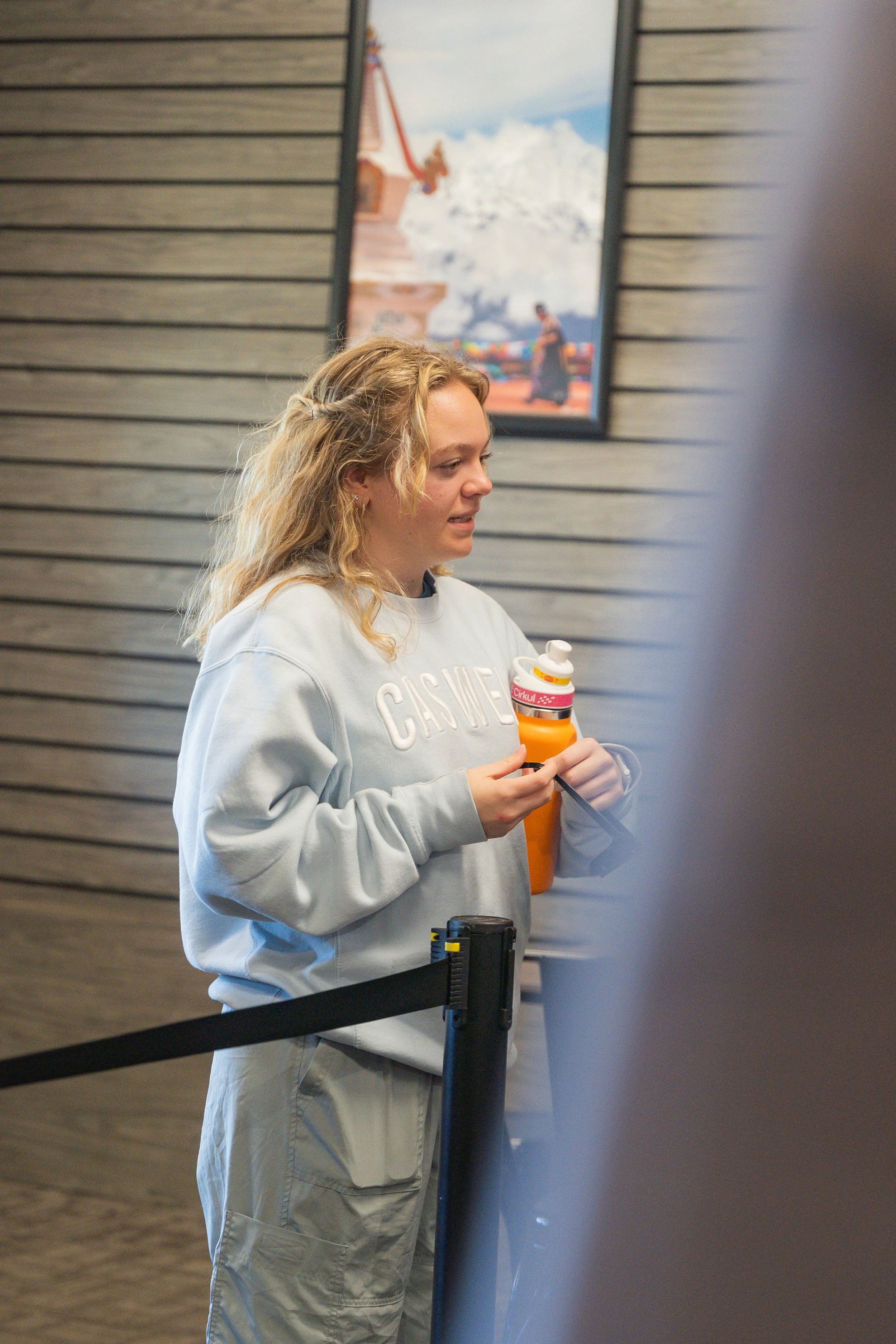 A woman with blonde curly hair, wearing a light grey sweatshirt and cargo pants, holding an orange bottled beverage in her hand, standing behind a black barrier in an indoor setting.