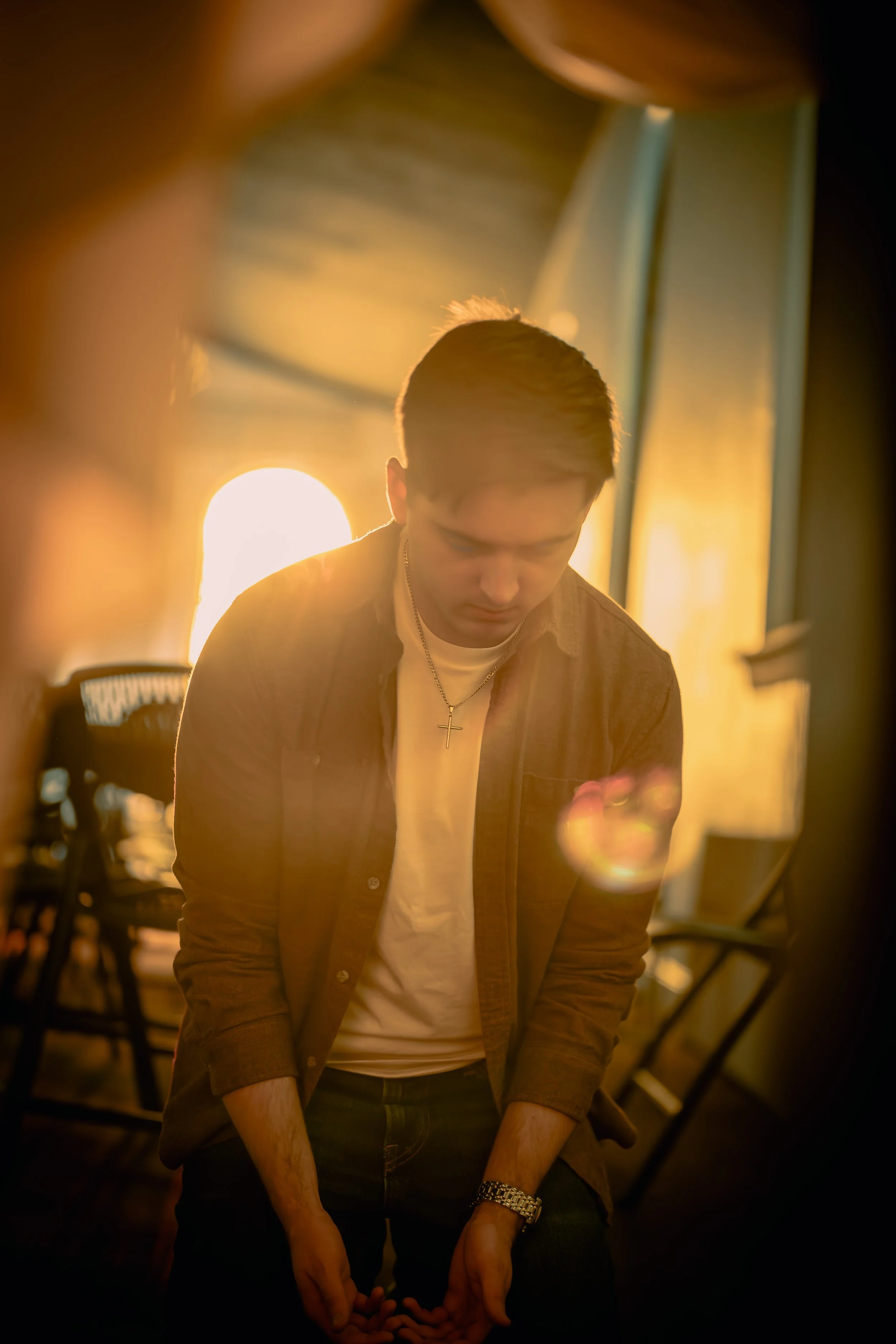 A young man with short brown hair, wearing a cross necklace, a white T-shirt, and a brown jacket, is looking downward with a serious expression. The background features warm, golden sunlight streaming through windows, creating a glow around him, with