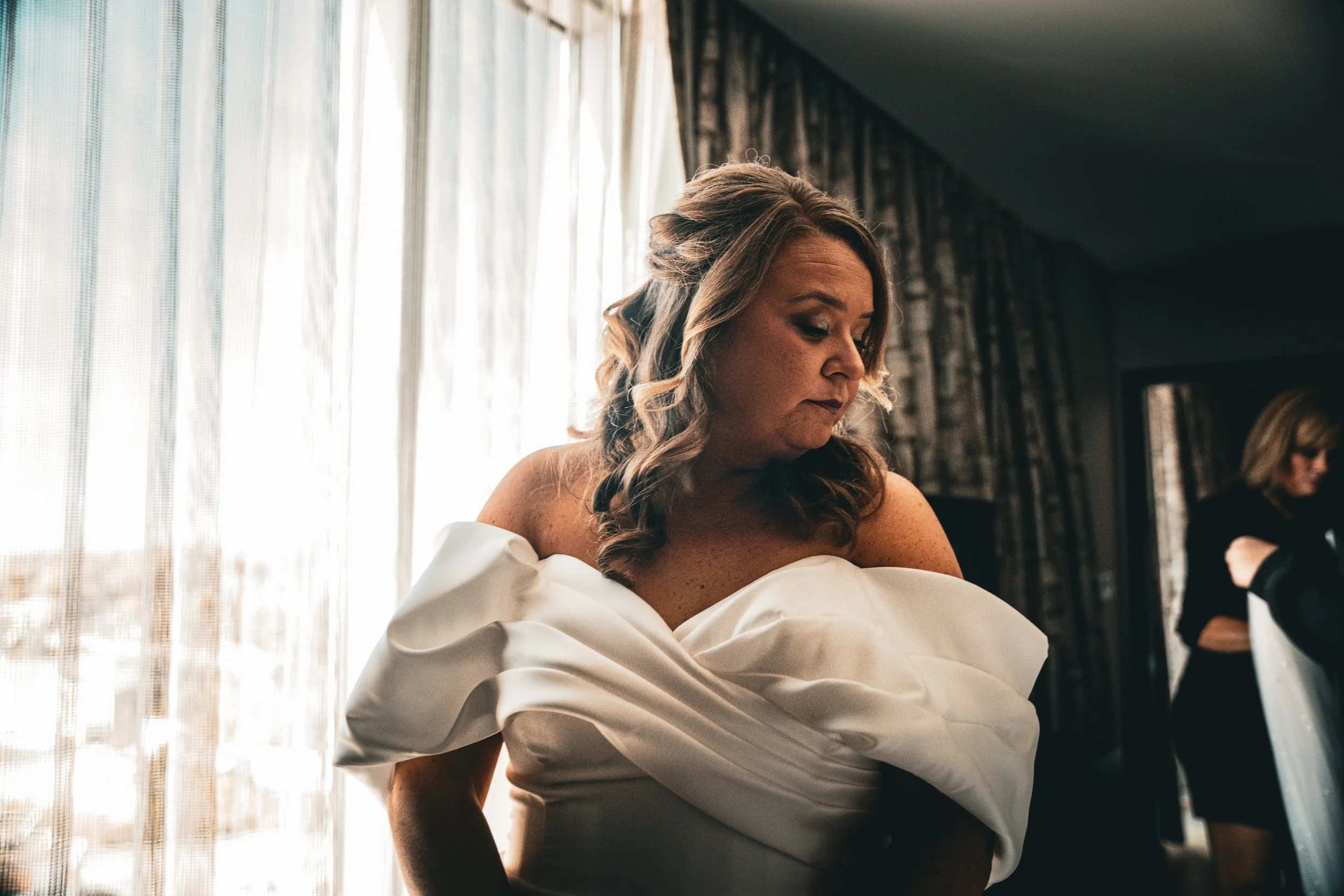 A woman with light skin and wavy, shoulder-length hair, wearing a white off-shoulder dress, looking down with a soft expression in a room with large sheer curtains and patterned dark curtains in the background.