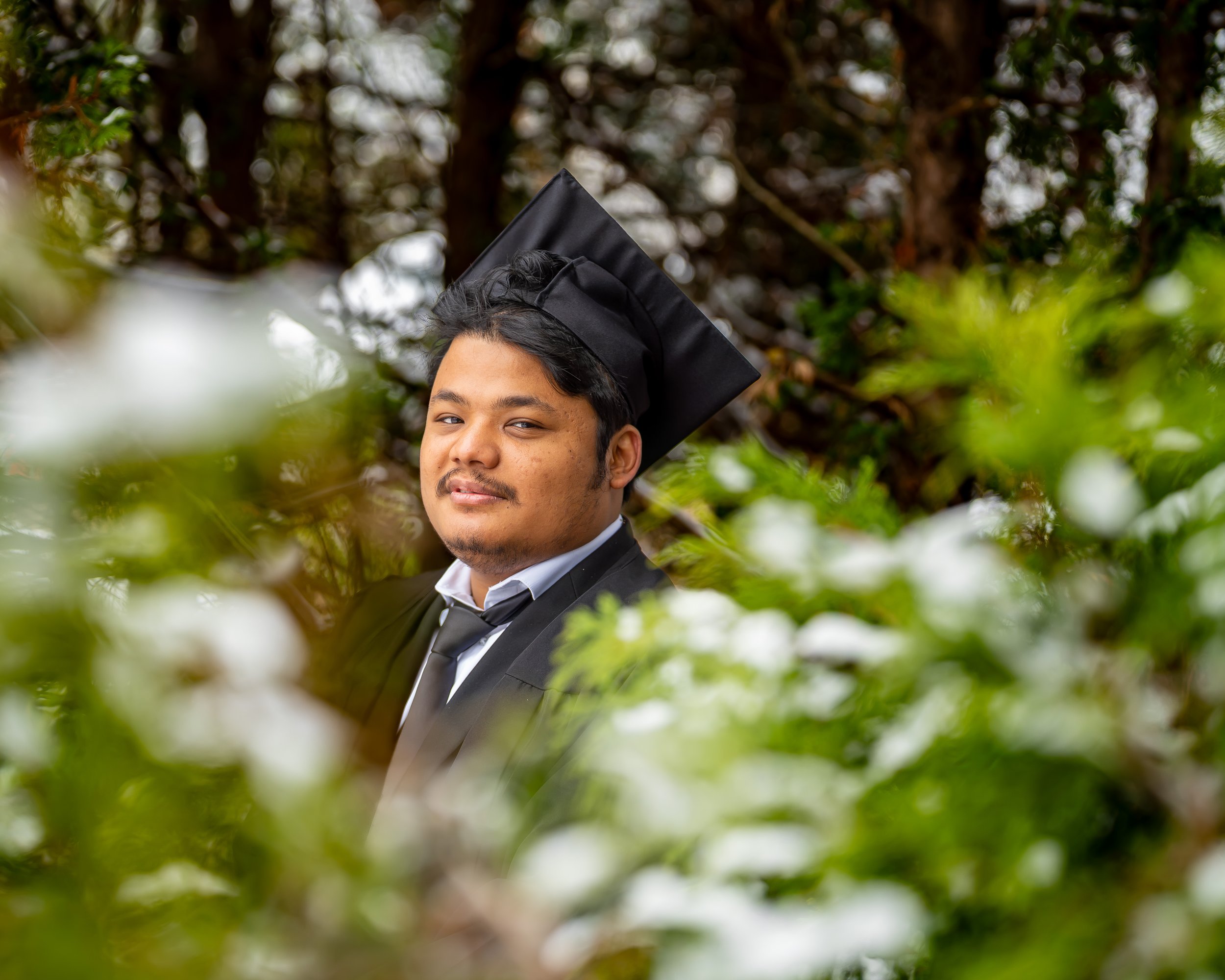 A young man in a graduation cap and gown poses outdoors among green foliage, with a background of trees and cloudy sky.
