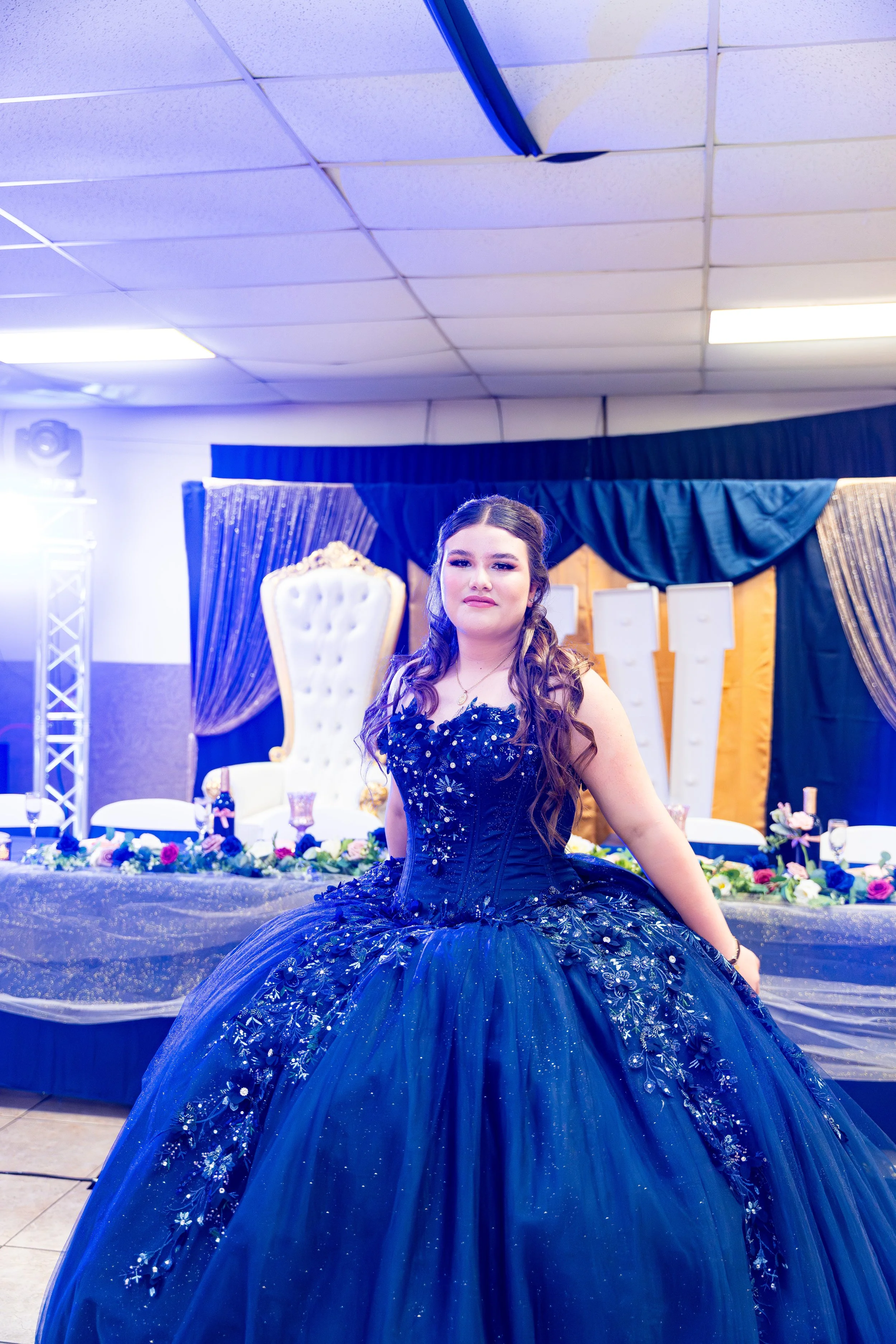 Young woman in an elegant dark blue ball gown with floral embroidery, seated at a decorated table in a banquet hall with a blue and gold theme.