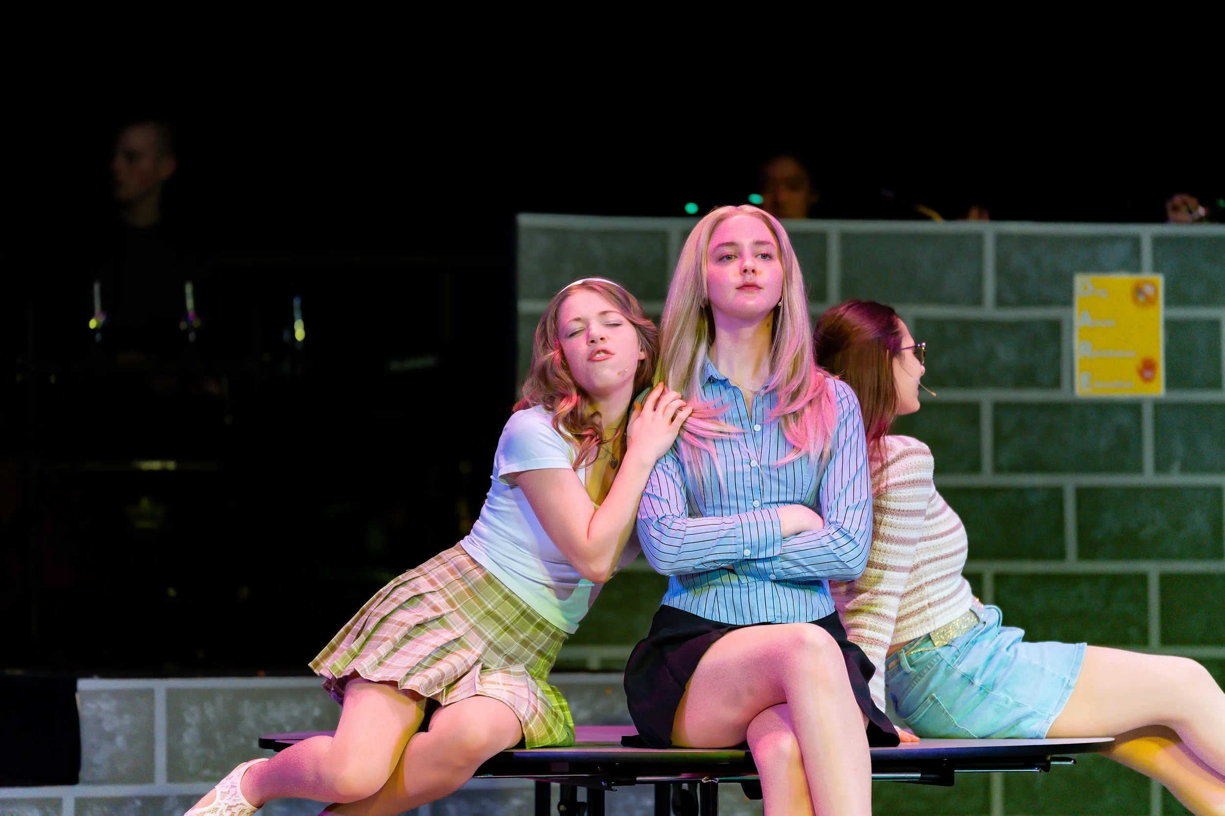 Four teenage girls on stage, one with eyes closed, one looking serious with arms crossed, sitting on a bench during a performance.