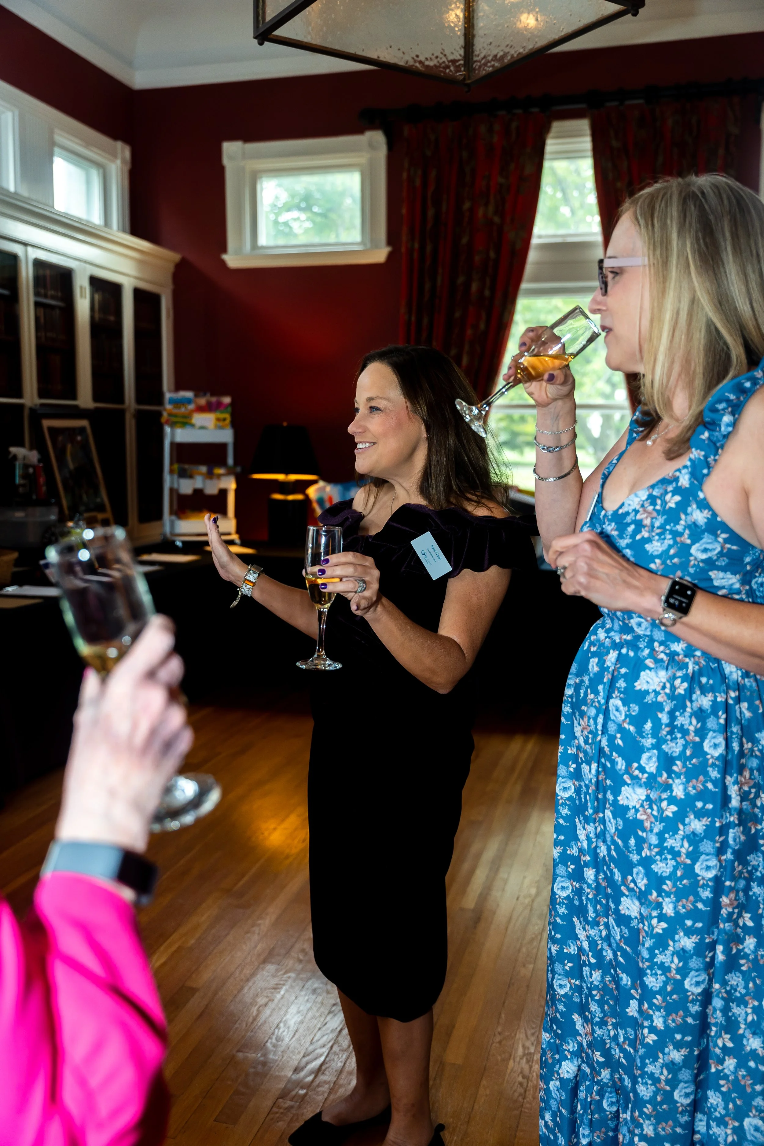 Women at a social gathering in a room with red walls and large windows, holding glasses of wine and smiling.
