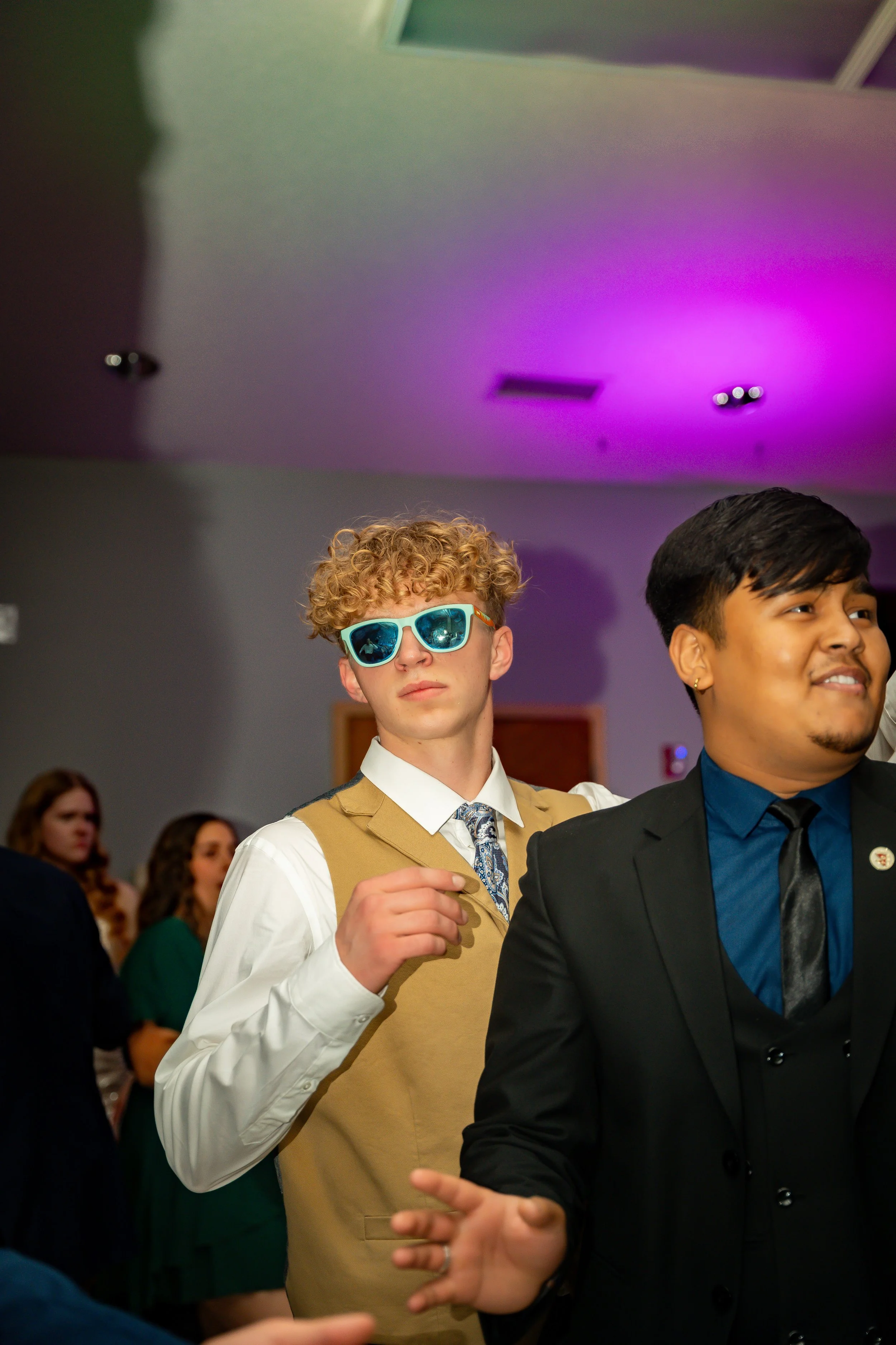 Two men dressed formally, one with curly hair wearing sunglasses and a vest, the other with straight hair wearing a dark suit, are dancing at an indoor event with colorful lighting.