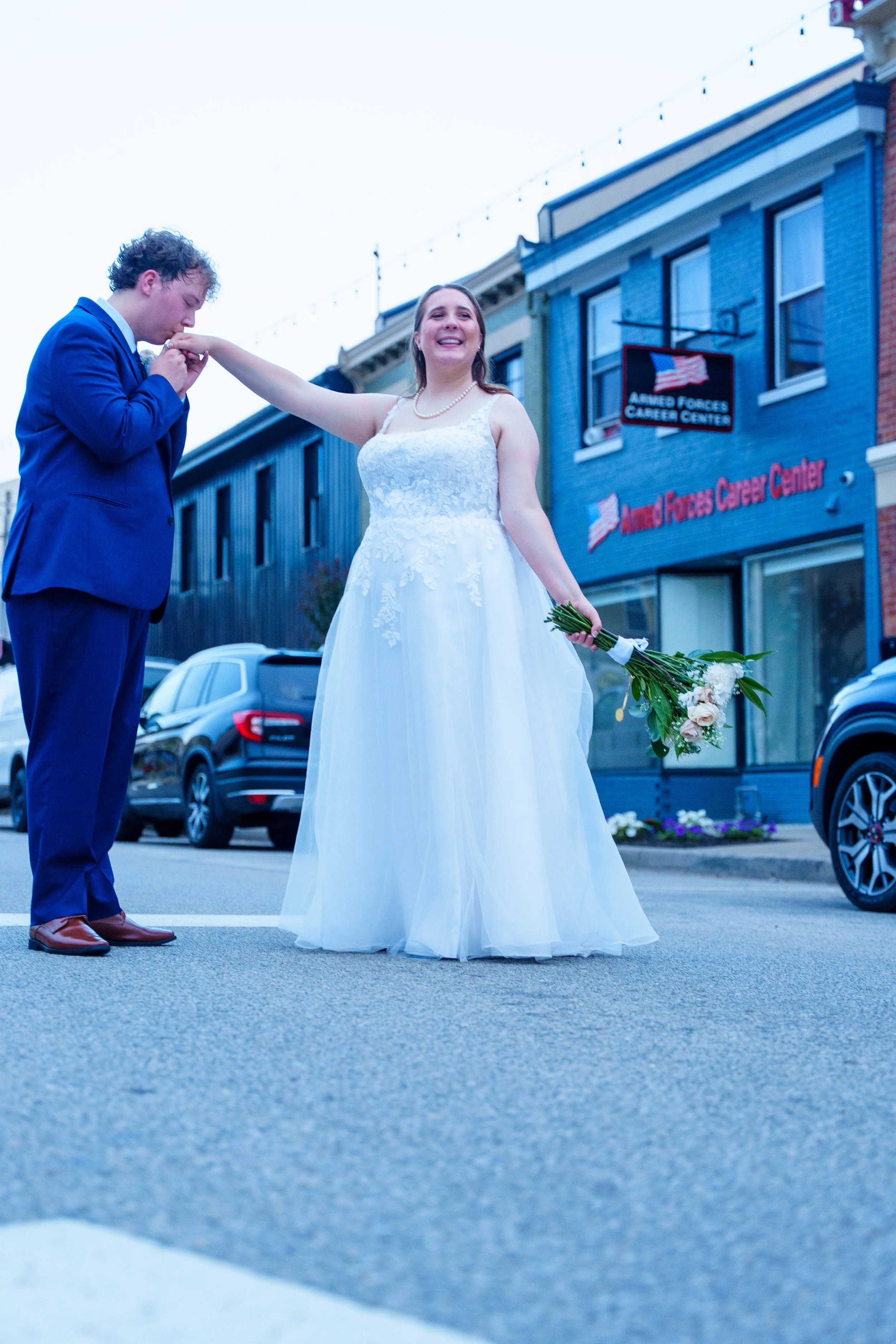 A bride in a white wedding dress smiling while holding a bouquet, kissing a groom in a blue suit on the hand in front of a building with a sign that reads 'Armed Forces Career Center.'