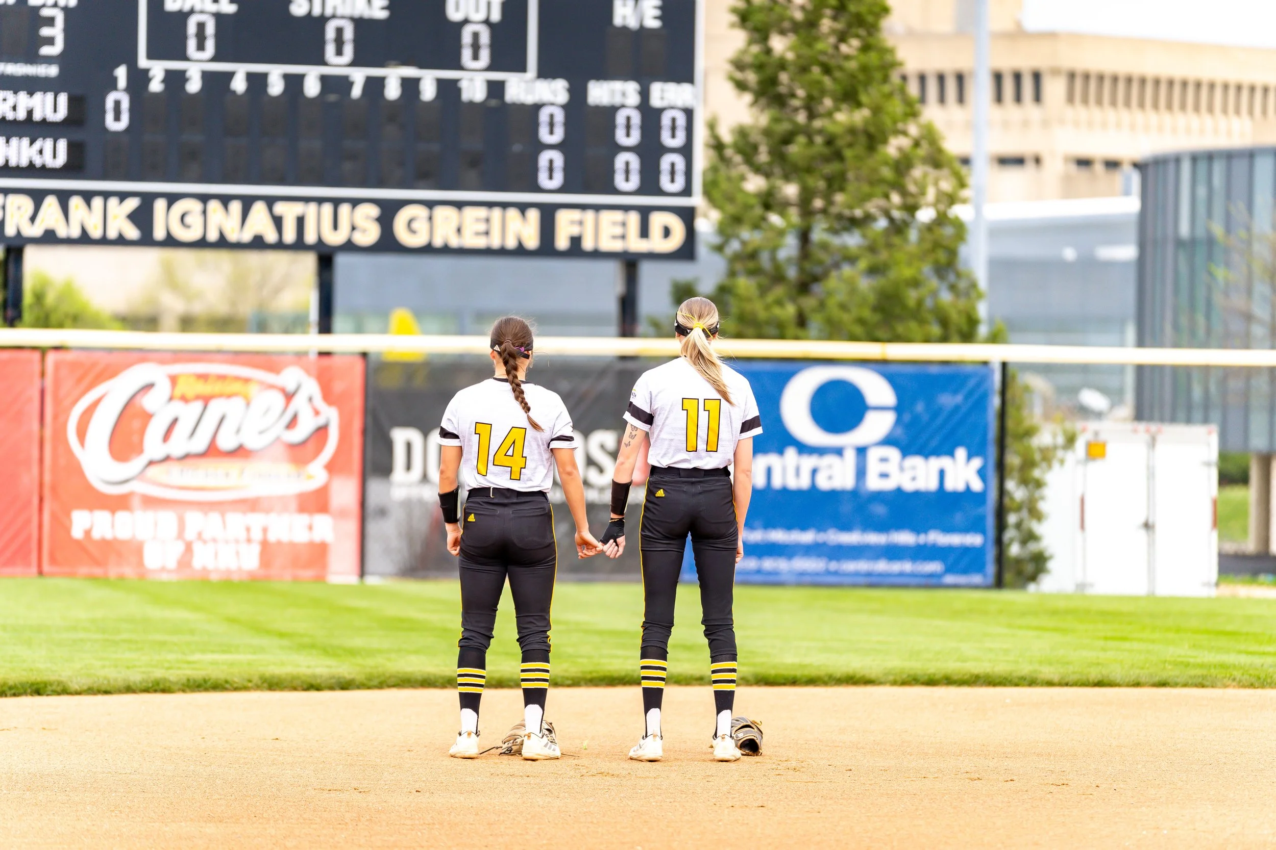Two female softball players holding hands on the field, wearing black and yellow uniforms with numbers 14 and 11, facing away from the camera, with a scoreboard and trees in the background.