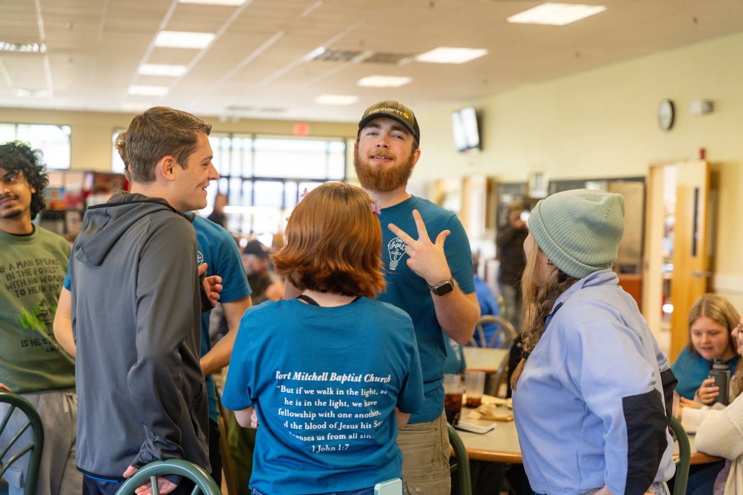 A group of five young people engaged in conversation inside a community room, with two women and three men, some wearing shirts with religious text, and the background showing large windows, a clock, and vending machines.