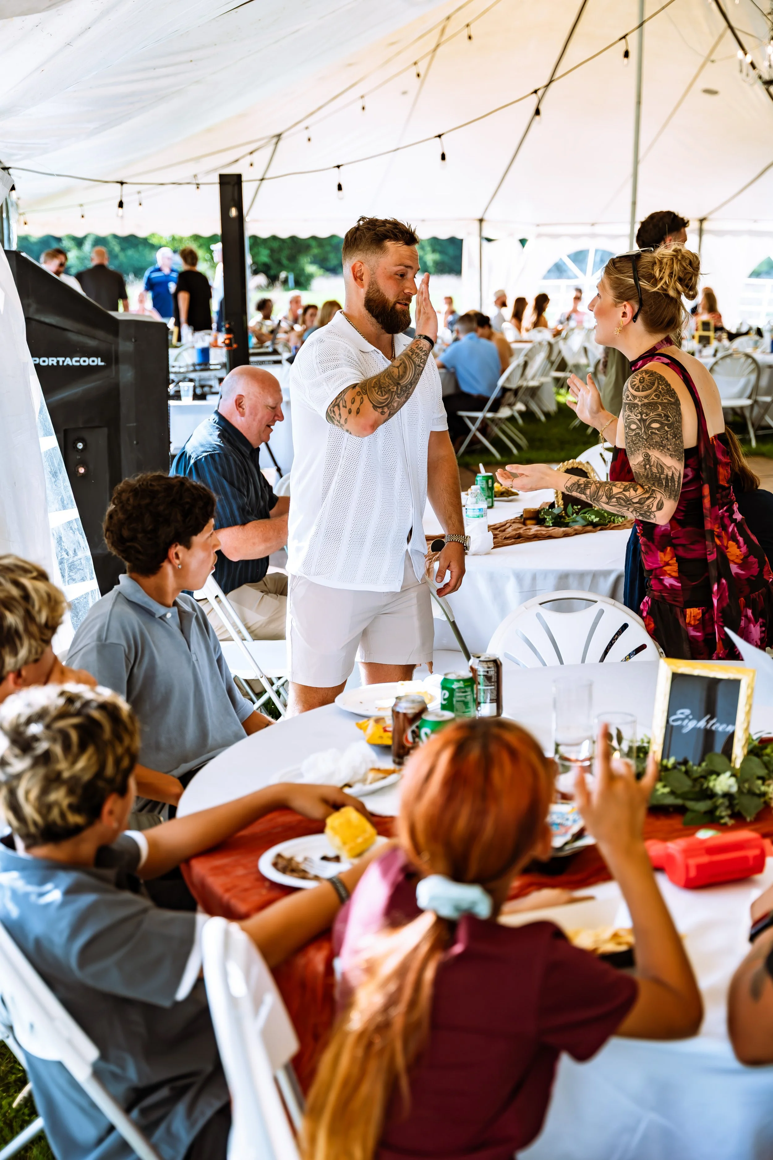 People at a large outdoor gathering under a tent, some sitting at tables, and a man with tattoos giving a high five to a woman with tattoos on her arm.
