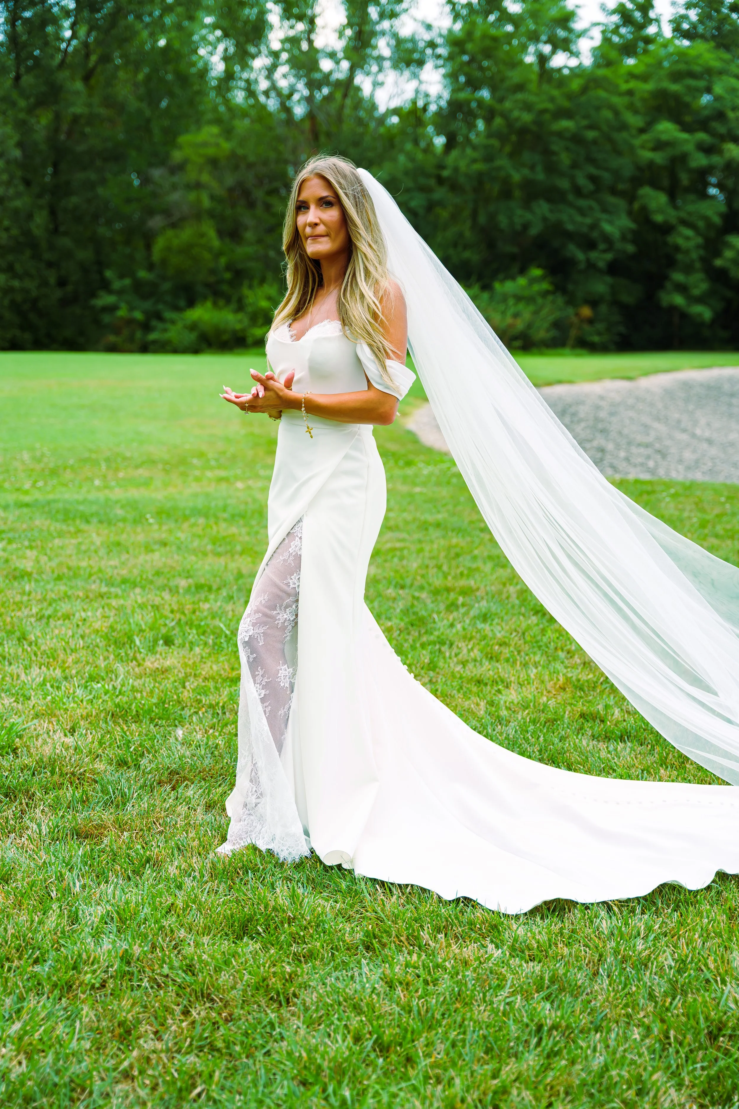 A bride in a white wedding dress standing on grass outdoors, with a green forest background and a gravel path. She has long blonde hair, a long veil, and is holding her hands together.