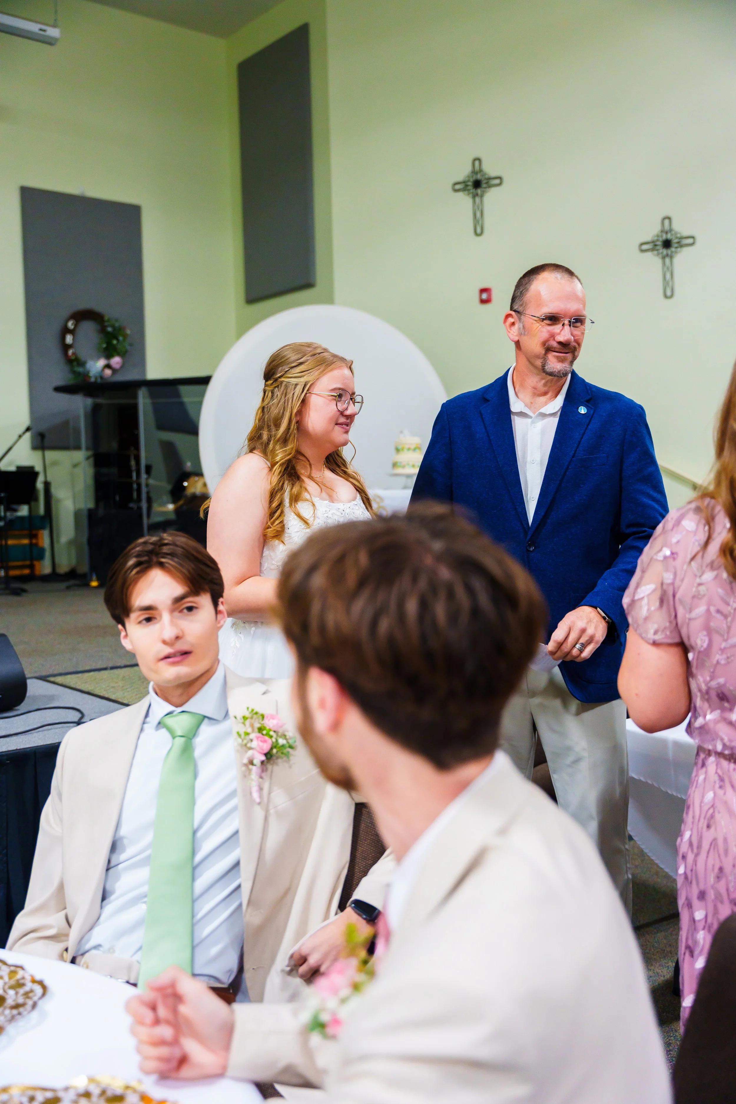 People dressed formally sitting at a table with a man and woman standing in the background, in a room decorated with crosses on the wall, possibly at a religious or wedding event.