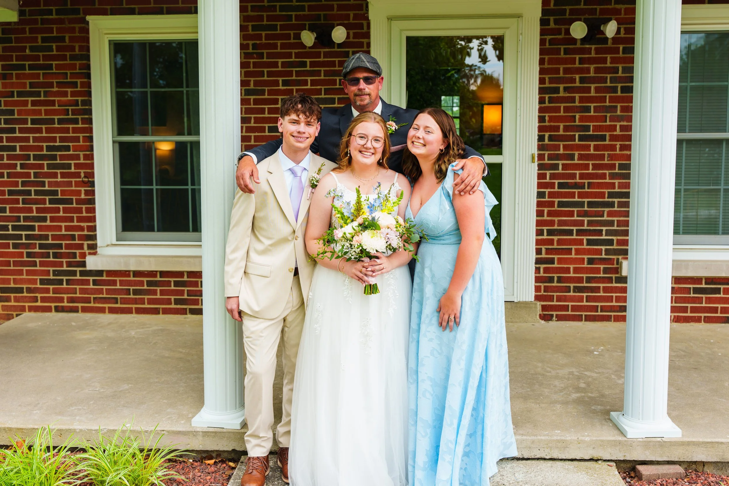 A group of five people standing outside a brick building, with a bride holding a bouquet of flowers, a man and woman on either side, and two other young individuals behind them, all smiling.