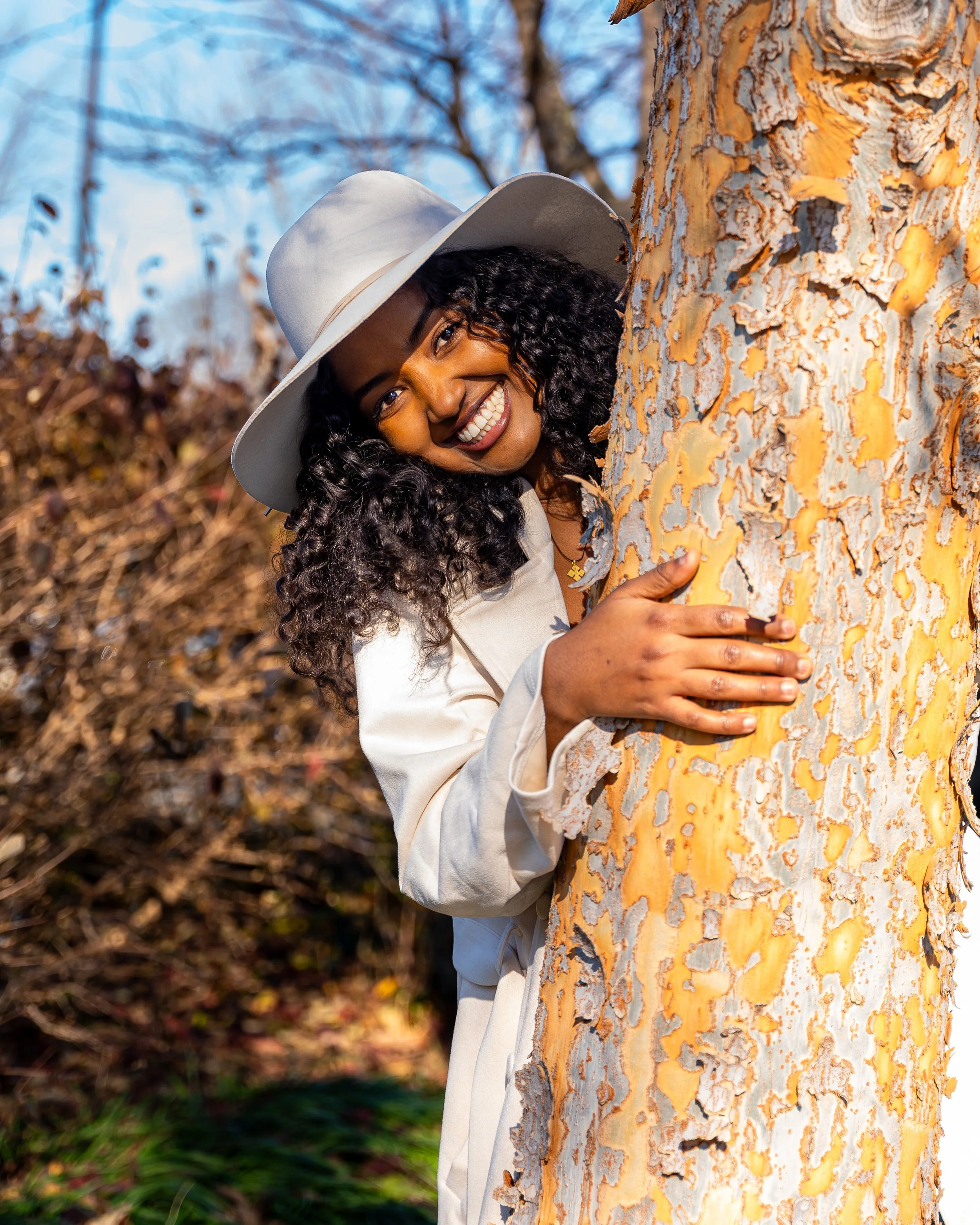 A woman with curly hair wearing a white hat and coat hugging a tree outdoors, smiling at the camera.