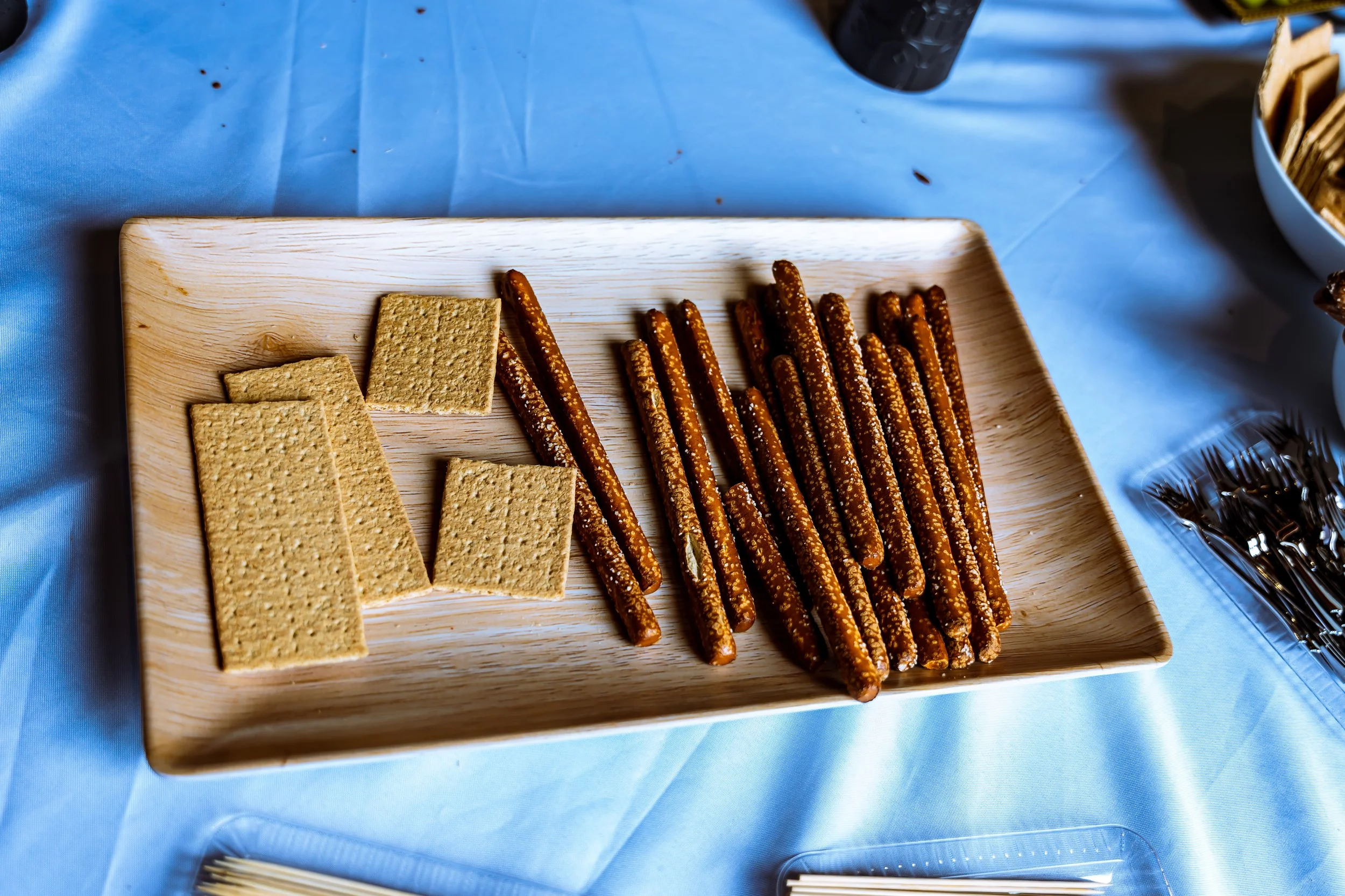 A wooden tray containing square graham crackers and pretzel sticks on a blue tablecloth.