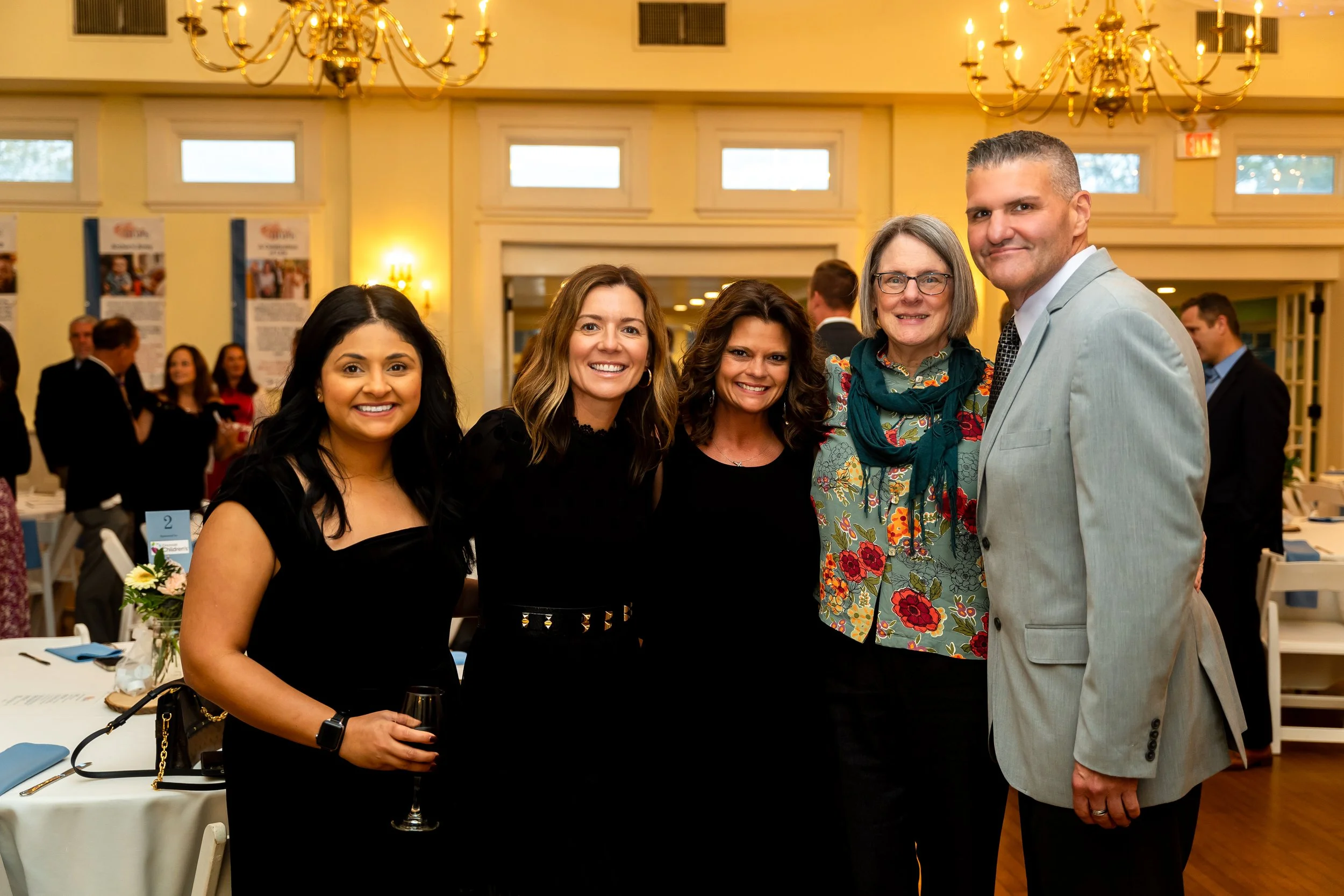Group of five smiling people, four women and one man, standing together at an indoor event with chandeliers and tables in the background.