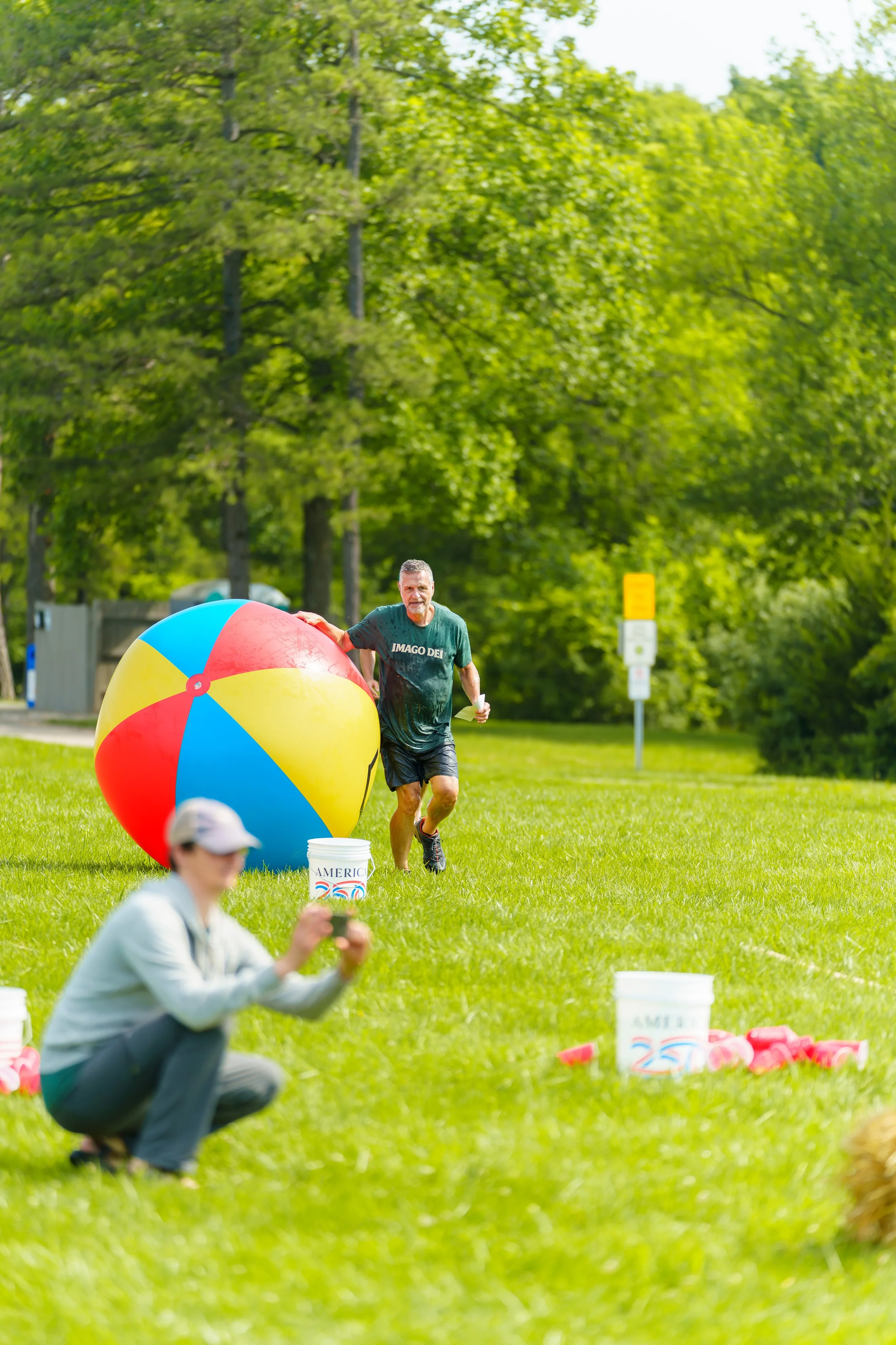A man smiling, wearing a dark green shirt and black shorts, holding a large colorful beach ball, outdoors on a grassy field with trees in the background, participating in a relay race or event.