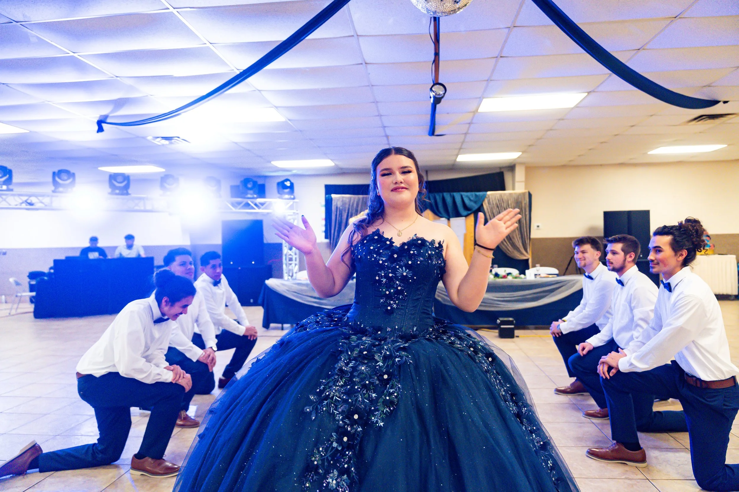 A young woman in a dark blue, embellished gown is dancing in the center of a group of men and women dressed in formal attire. The women are kneeling and the men are sitting in a line, in a decorated indoor hall with stage lights and a ceiling with ha