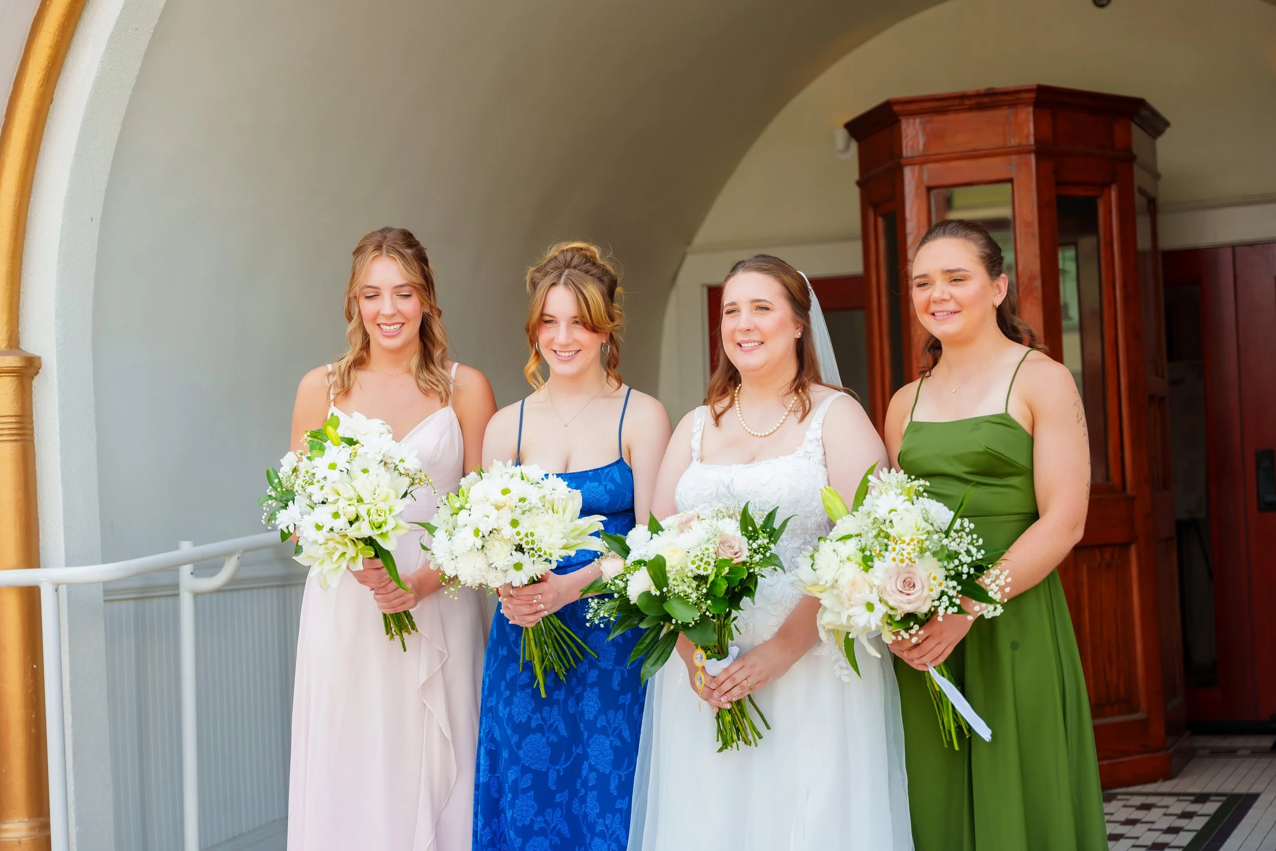Four women dressed in elegant dresses standing together, holding white and pink floral bouquets, smiling at a wedding or special event.