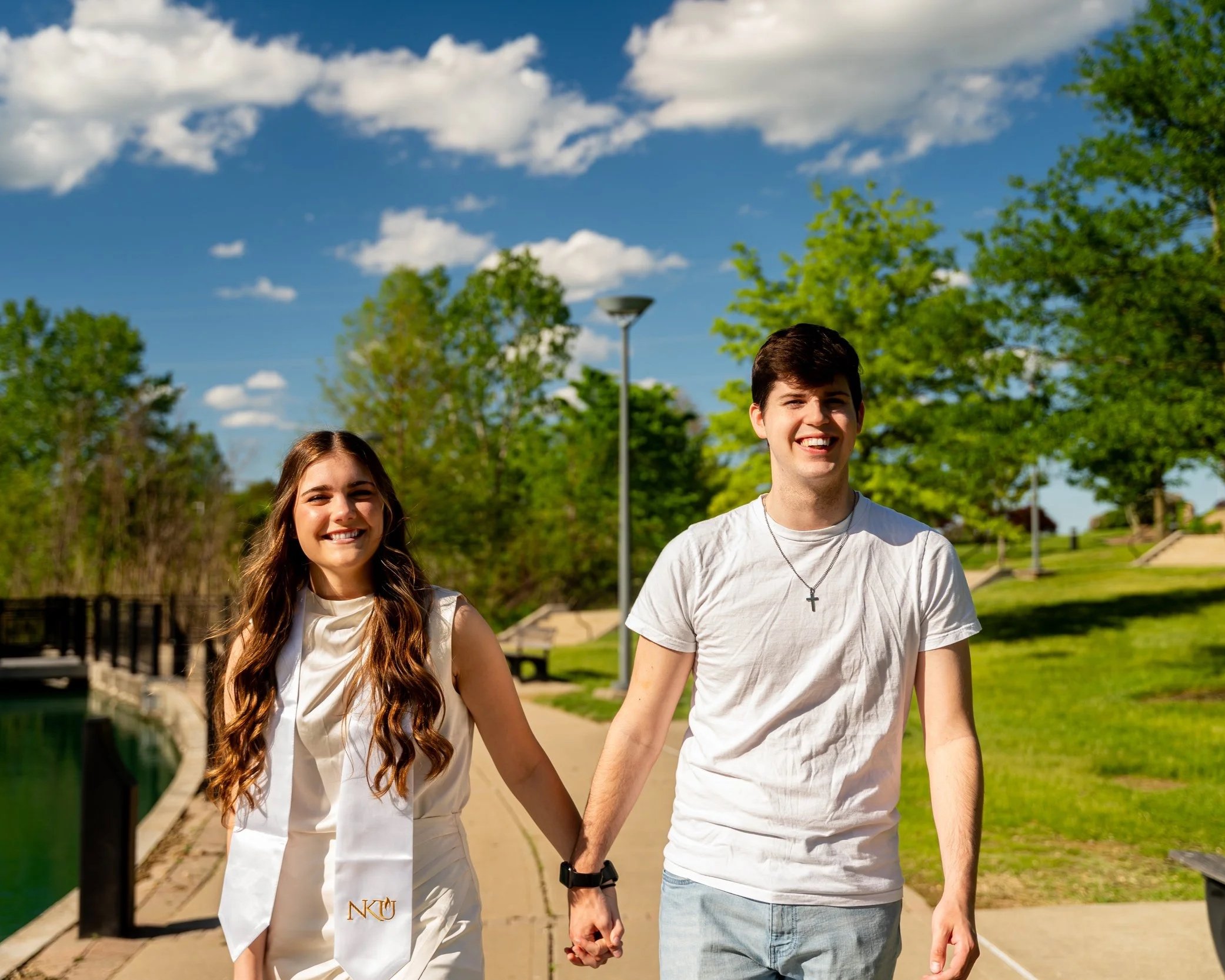 A young couple holding hands and walking on a paved path in a park on a sunny day, with green trees and a blue sky with white clouds in the background.