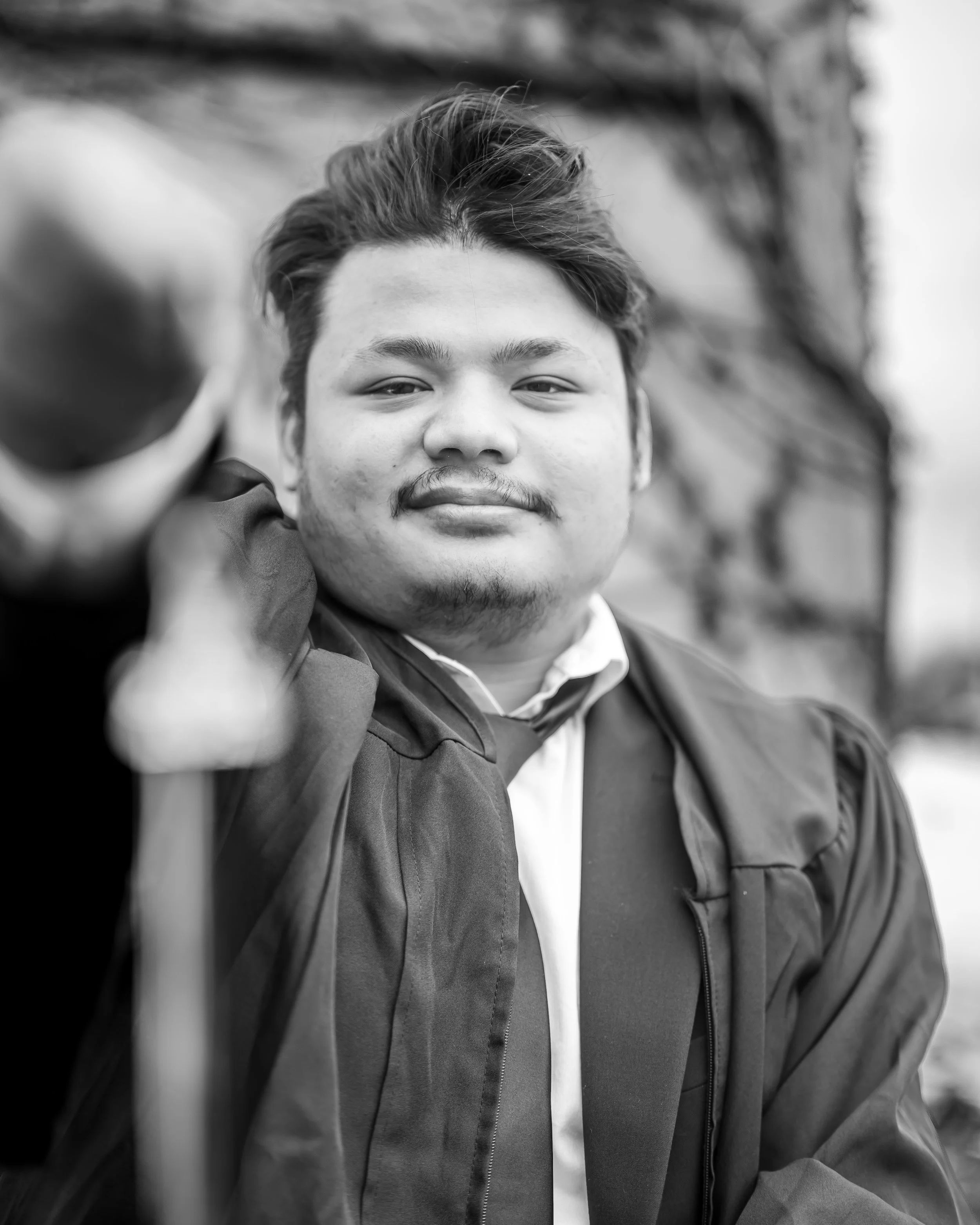 Black and white close-up portrait of a young man with styled hair, slight facial hair, and a confident expression, wearing a jacket and a white shirt, with a blurred outdoor background.