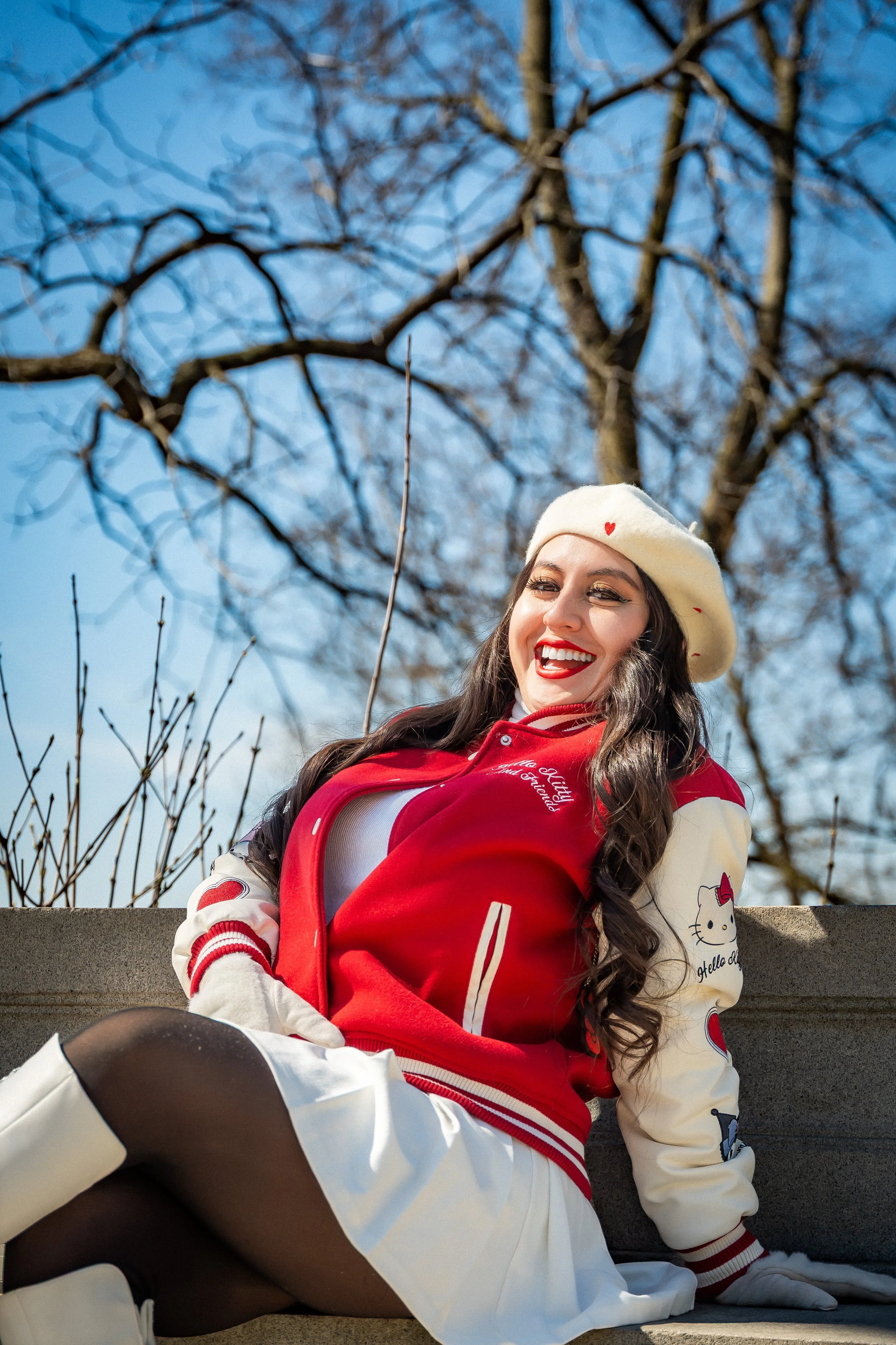 A woman sitting on a bench outdoors, smiling widely. She is wearing a white beret with red hearts, a red varsity jacket with white sleeves and Hello Kitty patches, a white skirt, black tights, and white knee-high boots. Bare tree branches and a clear