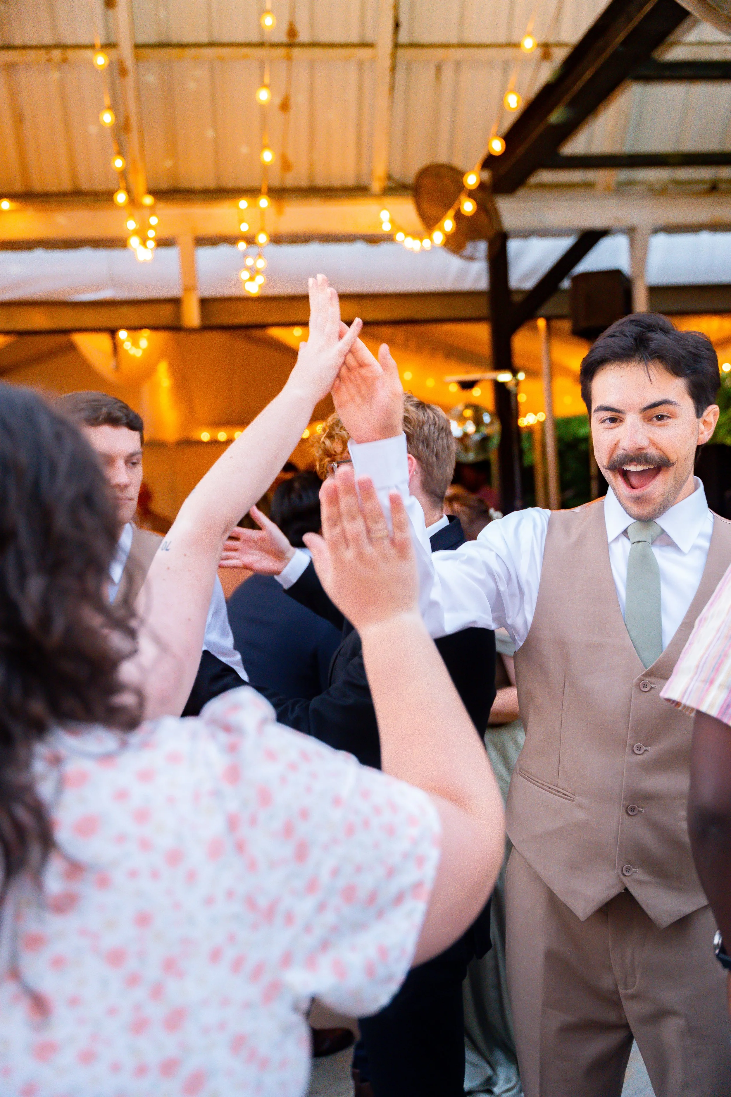 People celebrating at an indoor wedding reception, dancing and high-fiving under warm string lights.