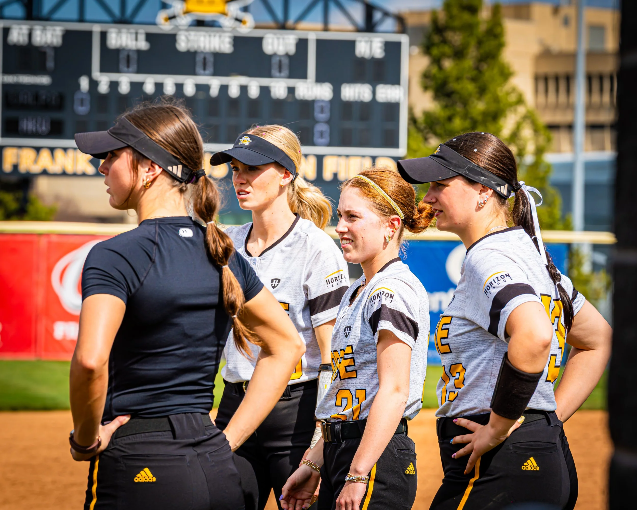 Group of female softball players standing in a line on the field, wearing team uniforms and visors, with a scoreboard and trees in the background.