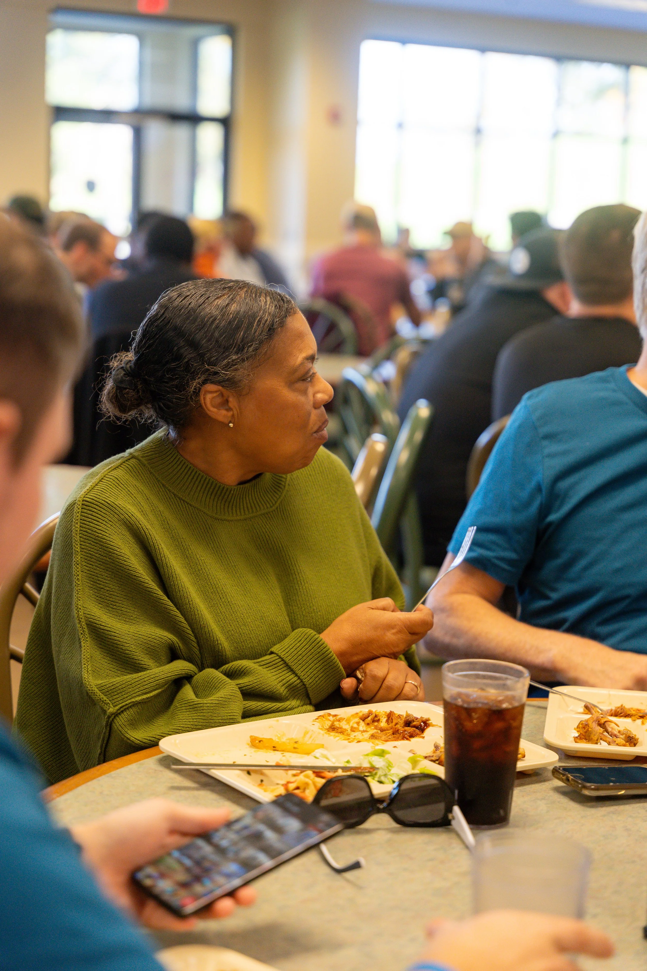 People sitting around a table at a social gathering or meeting, with food and drinks on the table, including a woman in a green sweater and a person in a blue shirt holding a phone.