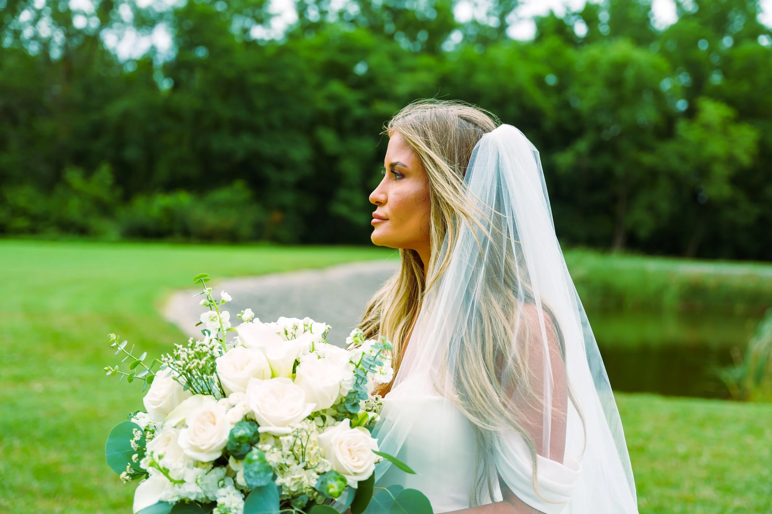 A bride with long blonde hair wearing a white wedding dress and veil is outdoors in a green park, holding a large bouquet of white roses and greenery.