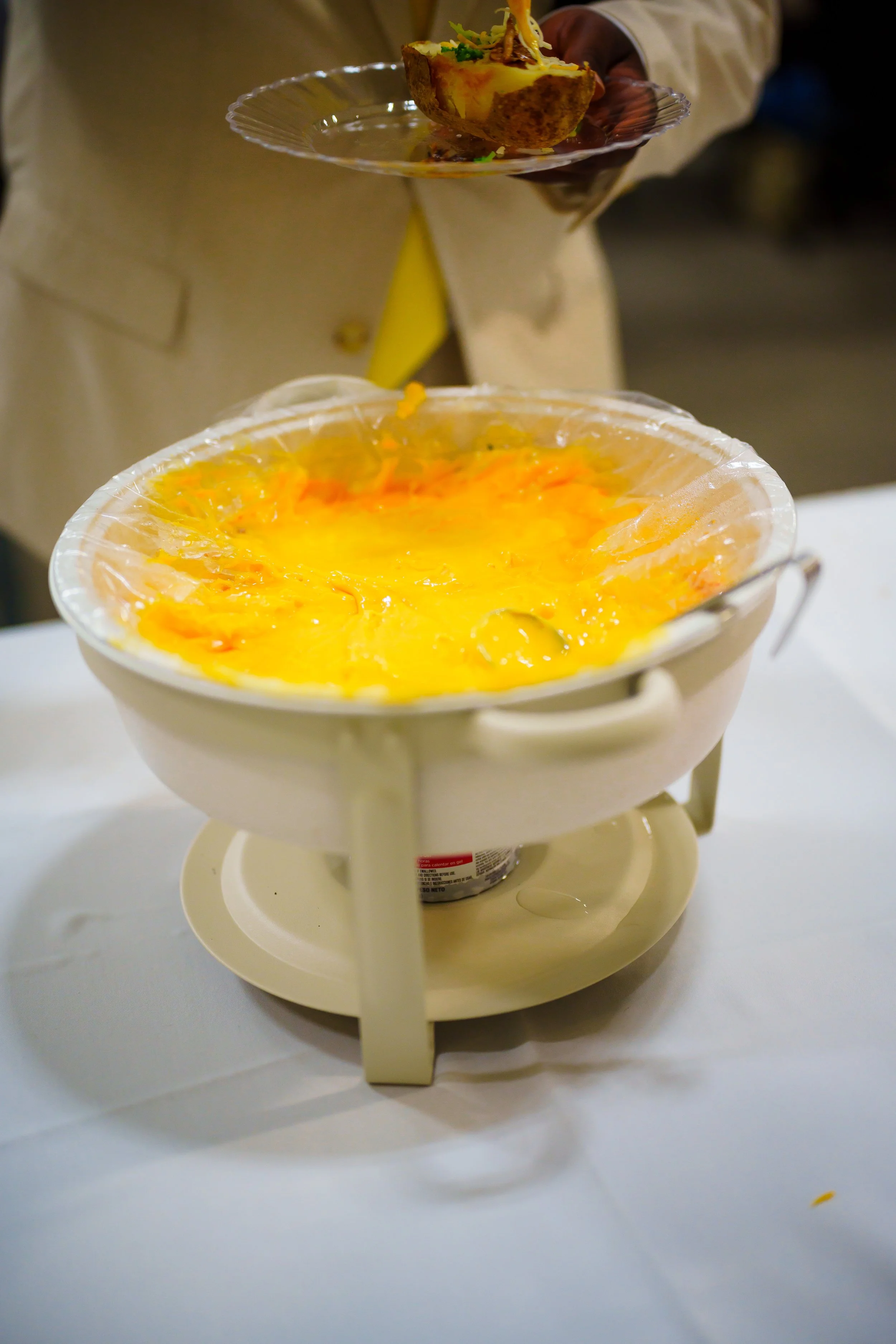 A vanilla or cheese fondue placed on a small stand with a burner underneath. The fondue is melting cheese with some bright orange and yellow colors. Someone is holding a plate with a bread bowl filled with cheese and toppings, ready to dip into the f