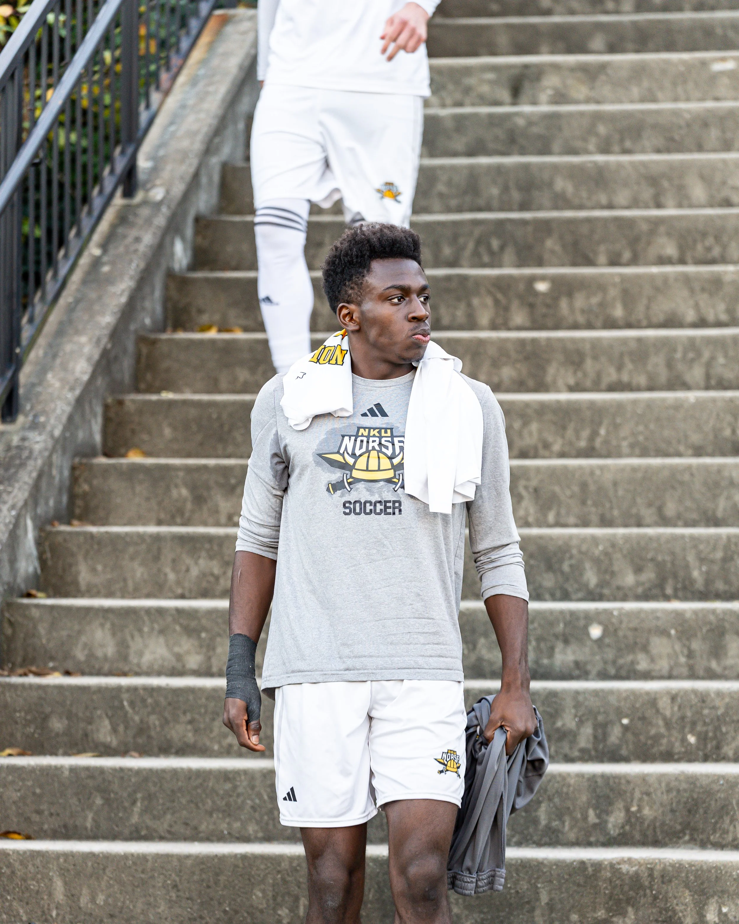 Young male soccer player walking down outdoor concrete stairs, wearing a gray soccer jersey with 'Norse' and soccer graphics, white shorts, and a white towel around his neck.