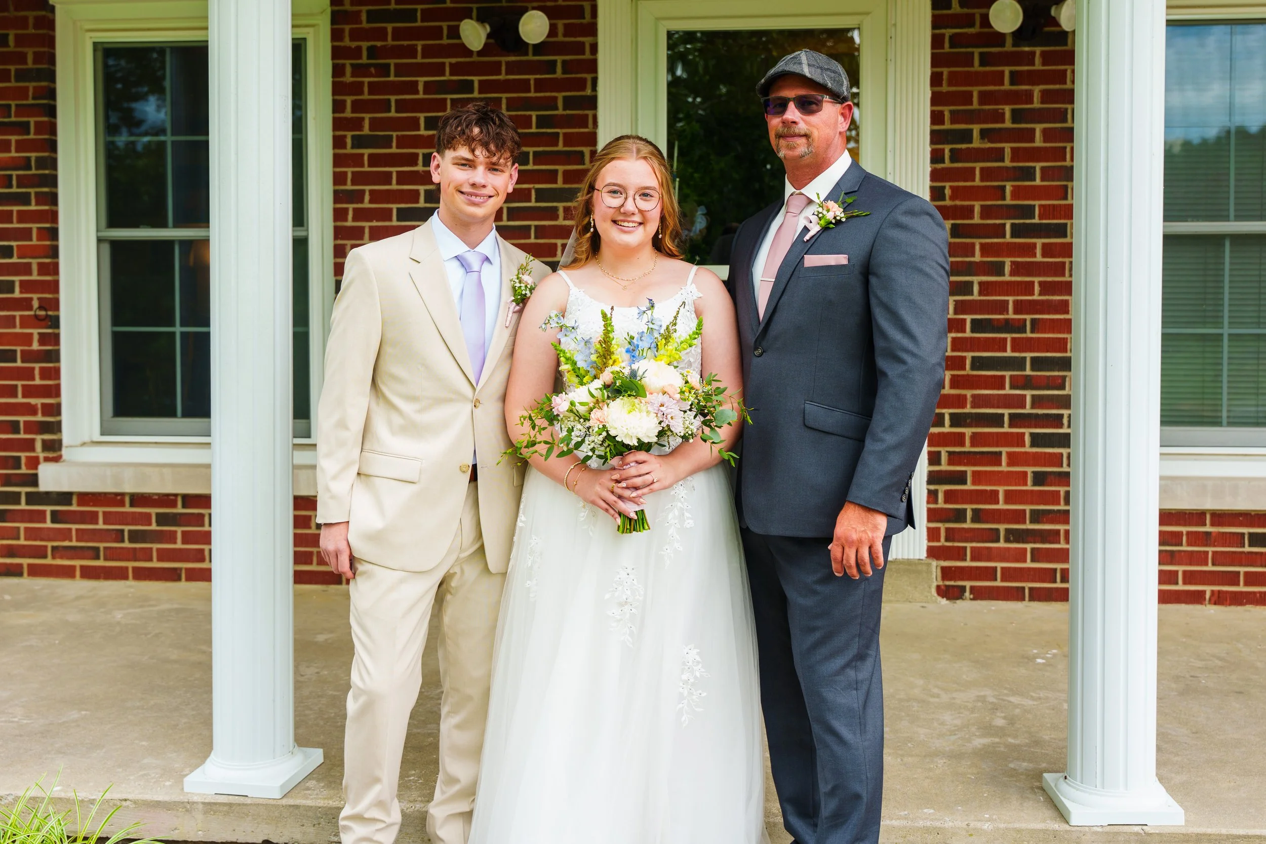 A wedding couple with a man and woman standing outdoors in front of a brick house, holding a bouquet of flowers, under a white porch structure.
