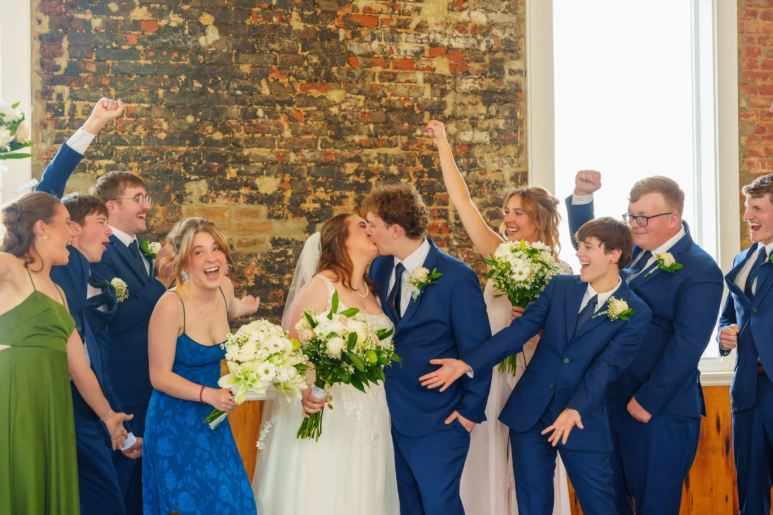 A group of people, including a bride and groom, celebrating at a wedding ceremony. The couple is kissing and holding hands, surrounded by friends and family who are cheering and raising their fists in joy, inside a room with a brick wall background.