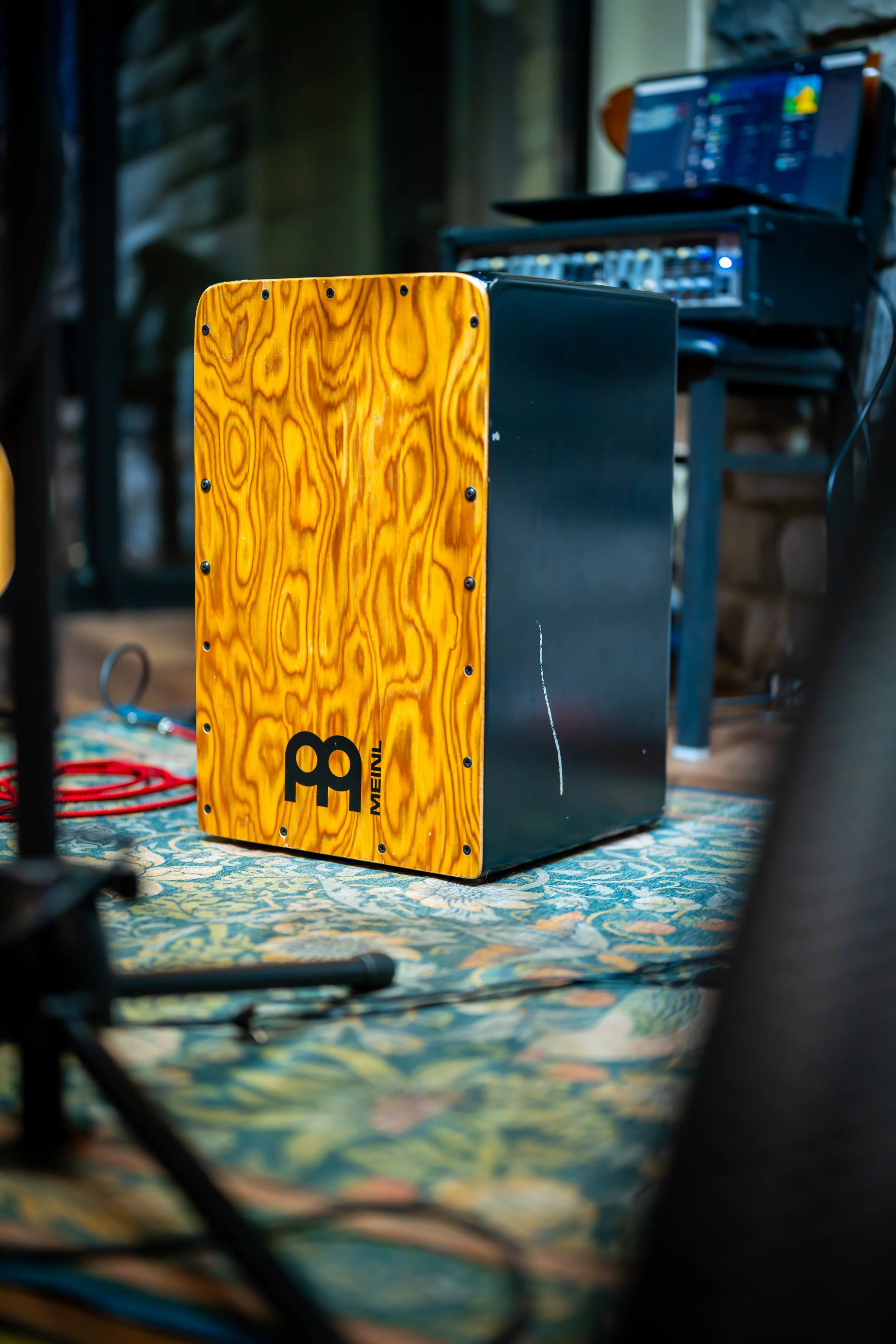 Close-up of a yellow wooden cajón drum on a patterned carpet with audio equipment in the background, in a dimly lit room.