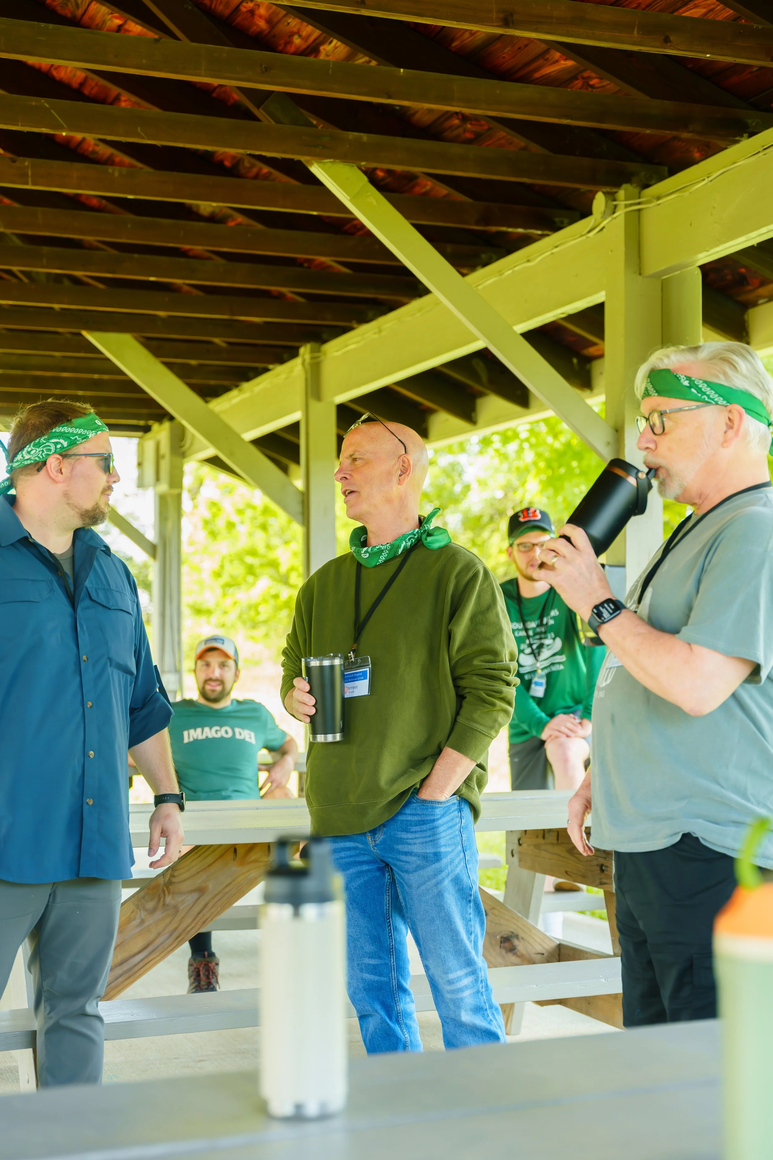 Group of people gathered outdoors under a pavilion, wearing casual clothing and green bandanas, engaged in conversation with trees in the background.