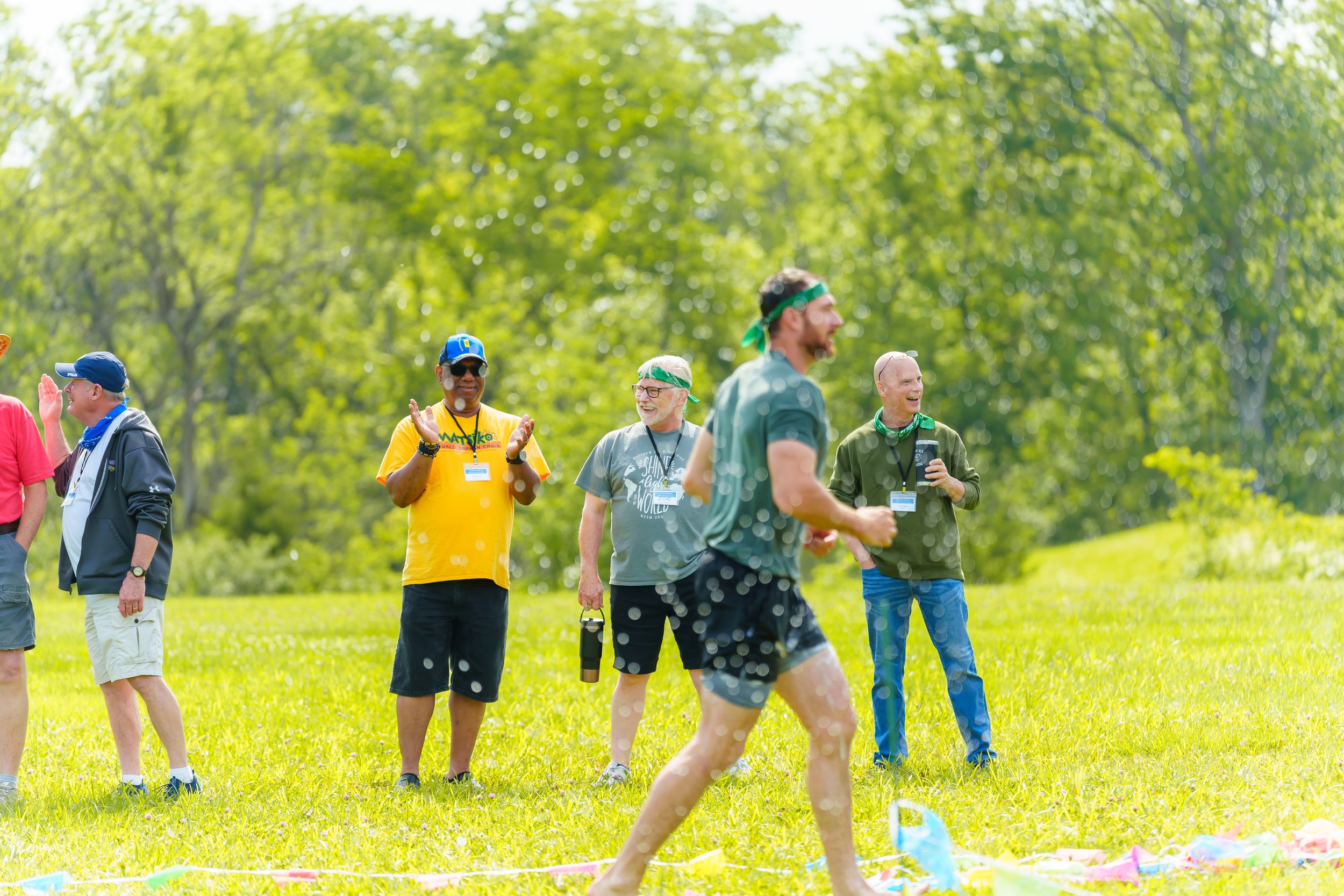 Group of people outdoors celebrating at a colorful event, with a young person in shorts running past. The group appears happy, some are clapping or smiling, and they are dressed casually with some wearing lanyards and headbands. There is greenery and