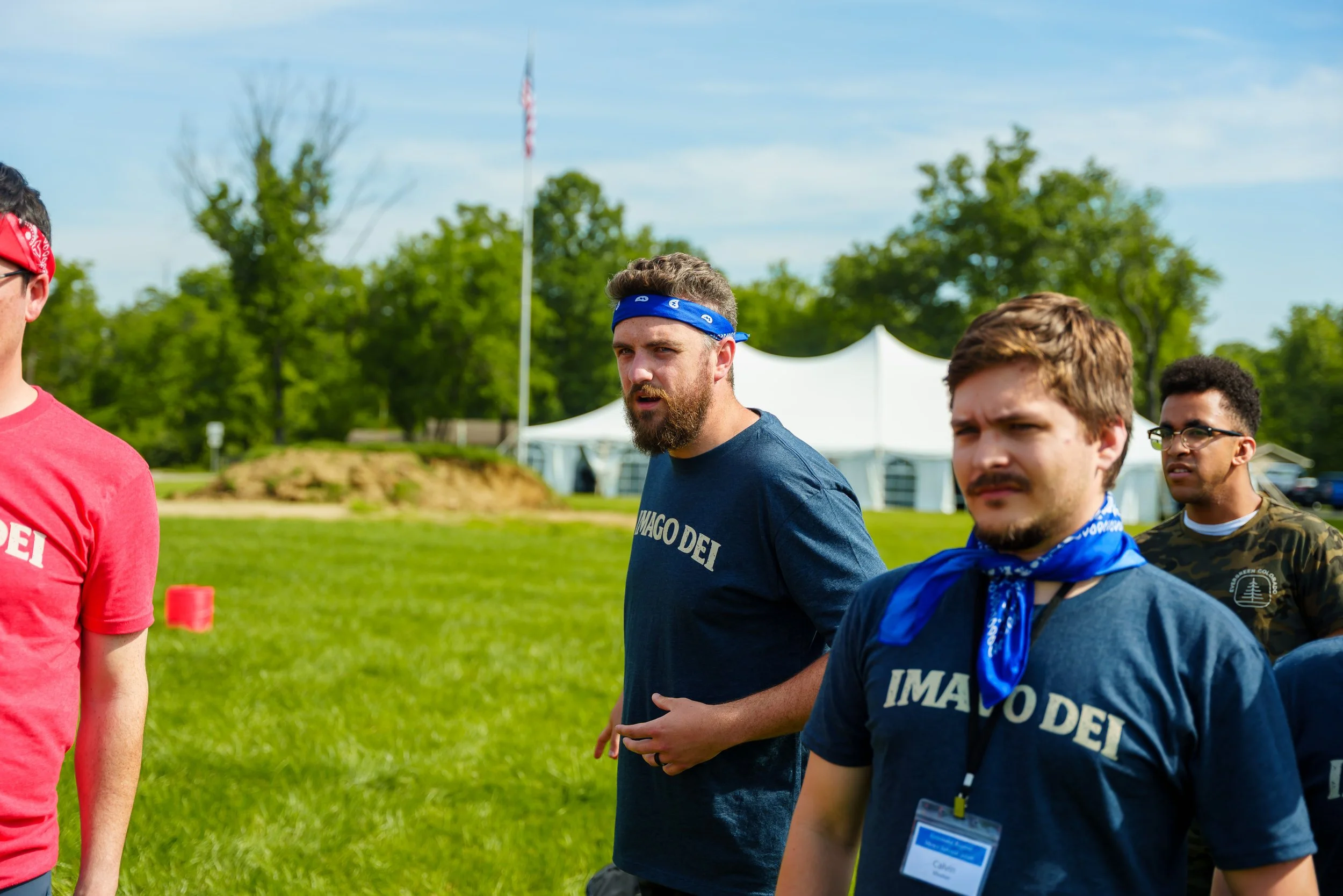 Group of young men standing outdoors during daytime, wearing patriotic bandanas and shirts, with green trees and a white tent in the background.