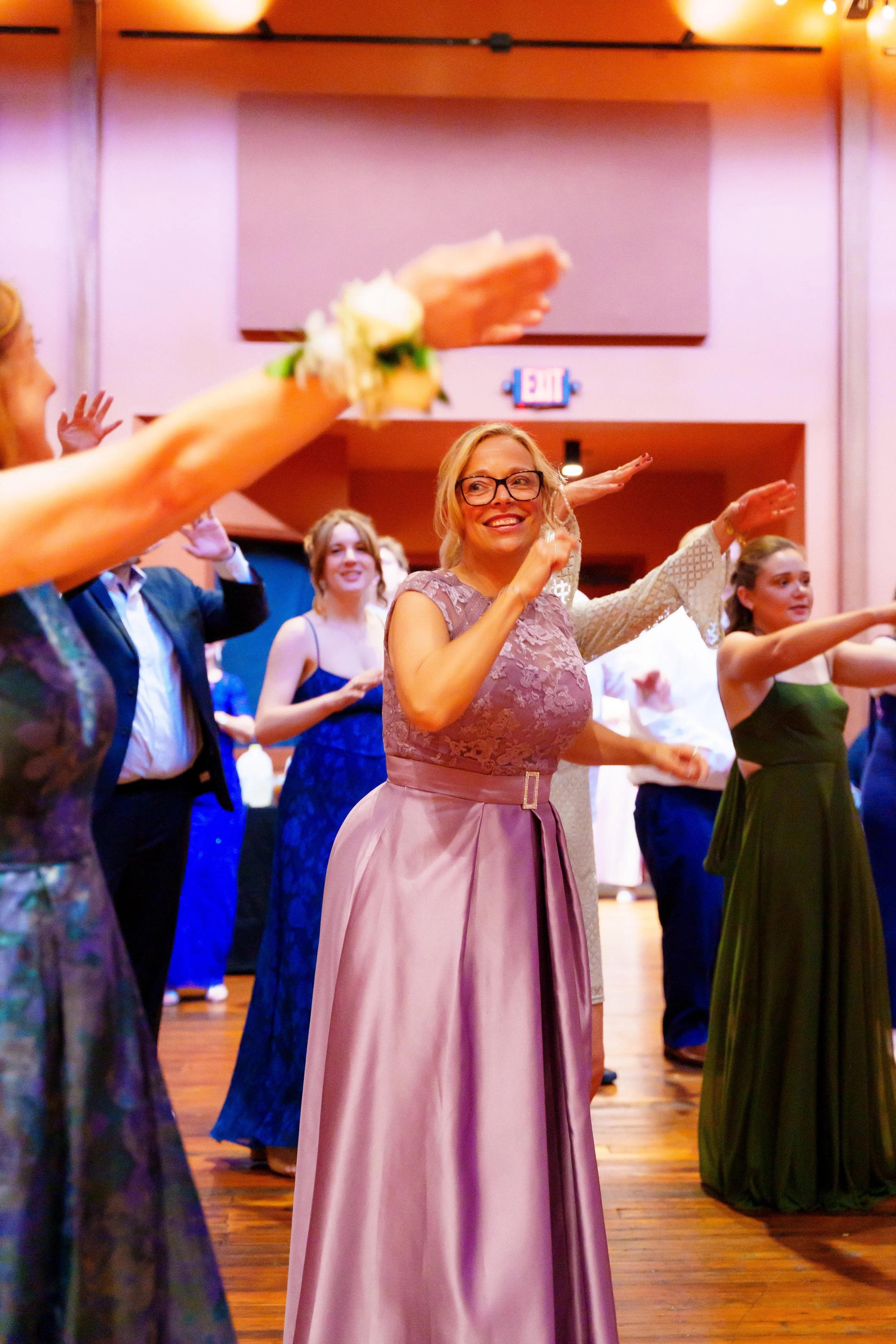 People dancing at a formal event or celebration in a hall, dressed in elegant attire, with one woman smiling and wearing glasses at the center of the image.
