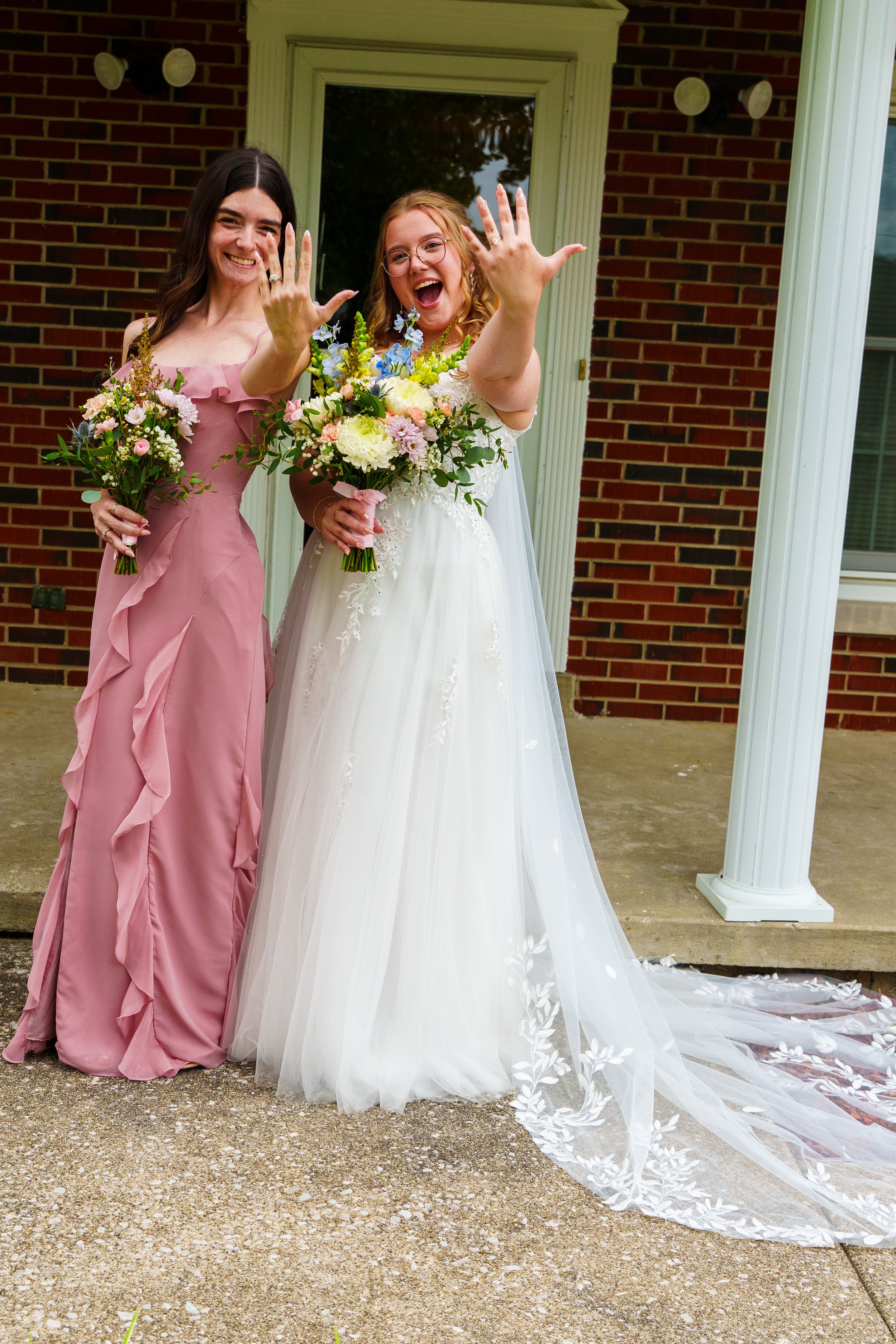 Two women celebrating a wedding, one in a white wedding dress holding a bouquet, and the other in a pink bridesmaid dress holding a smaller bouquet, standing outside in front of a brick house with a white column and a window.