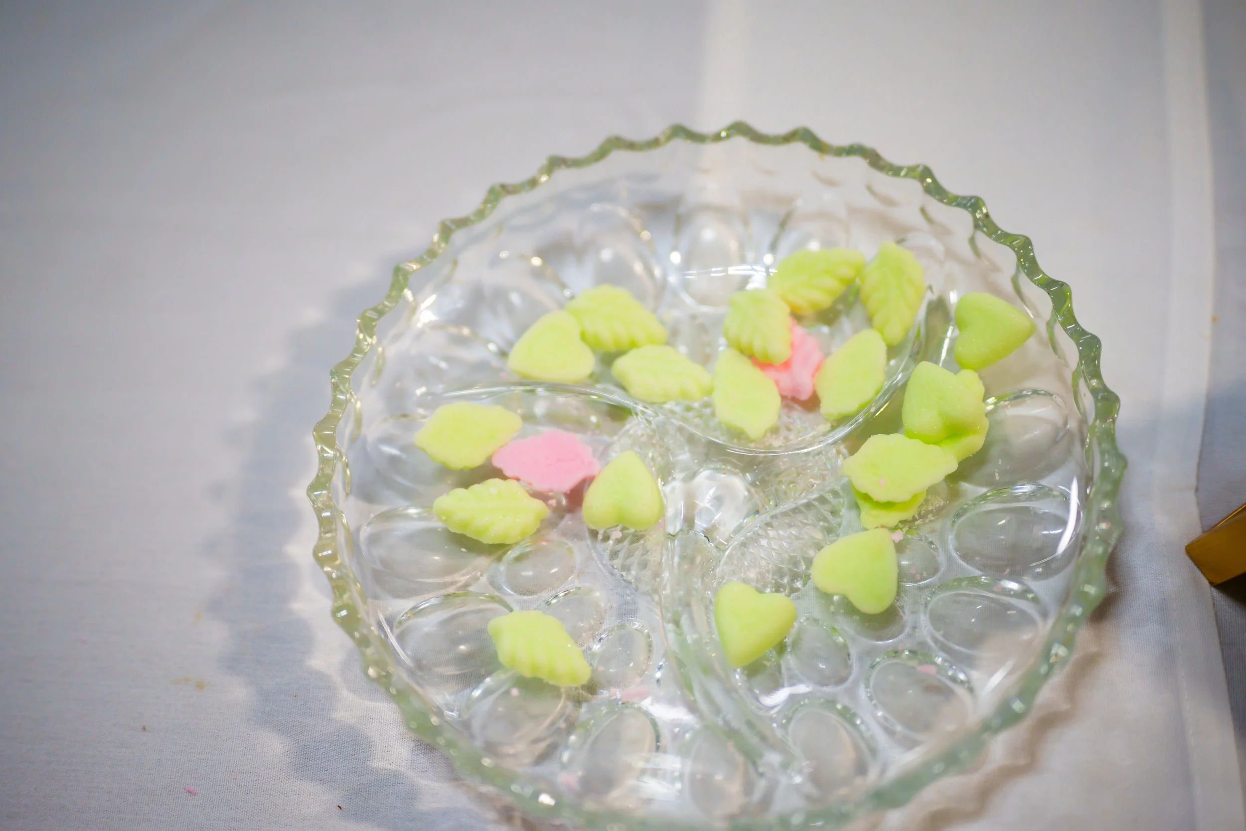 Decorative glass dish with pastel-colored candies, placed on a white surface with a pink item partially visible on the side.