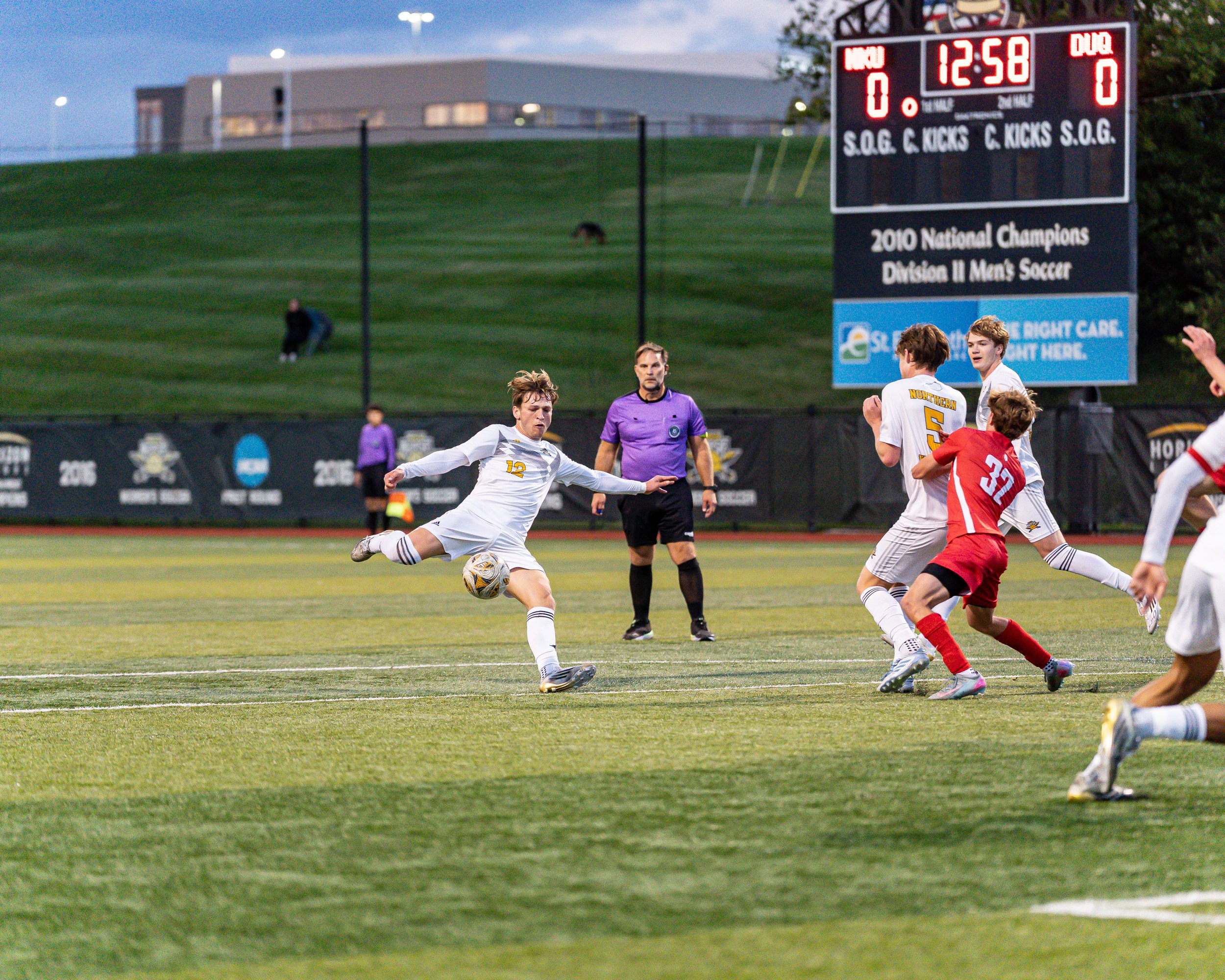 A soccer game with players in white and red uniforms on a grassy field, with a digital scoreboard showing 12:58 and a referee in purple observing.