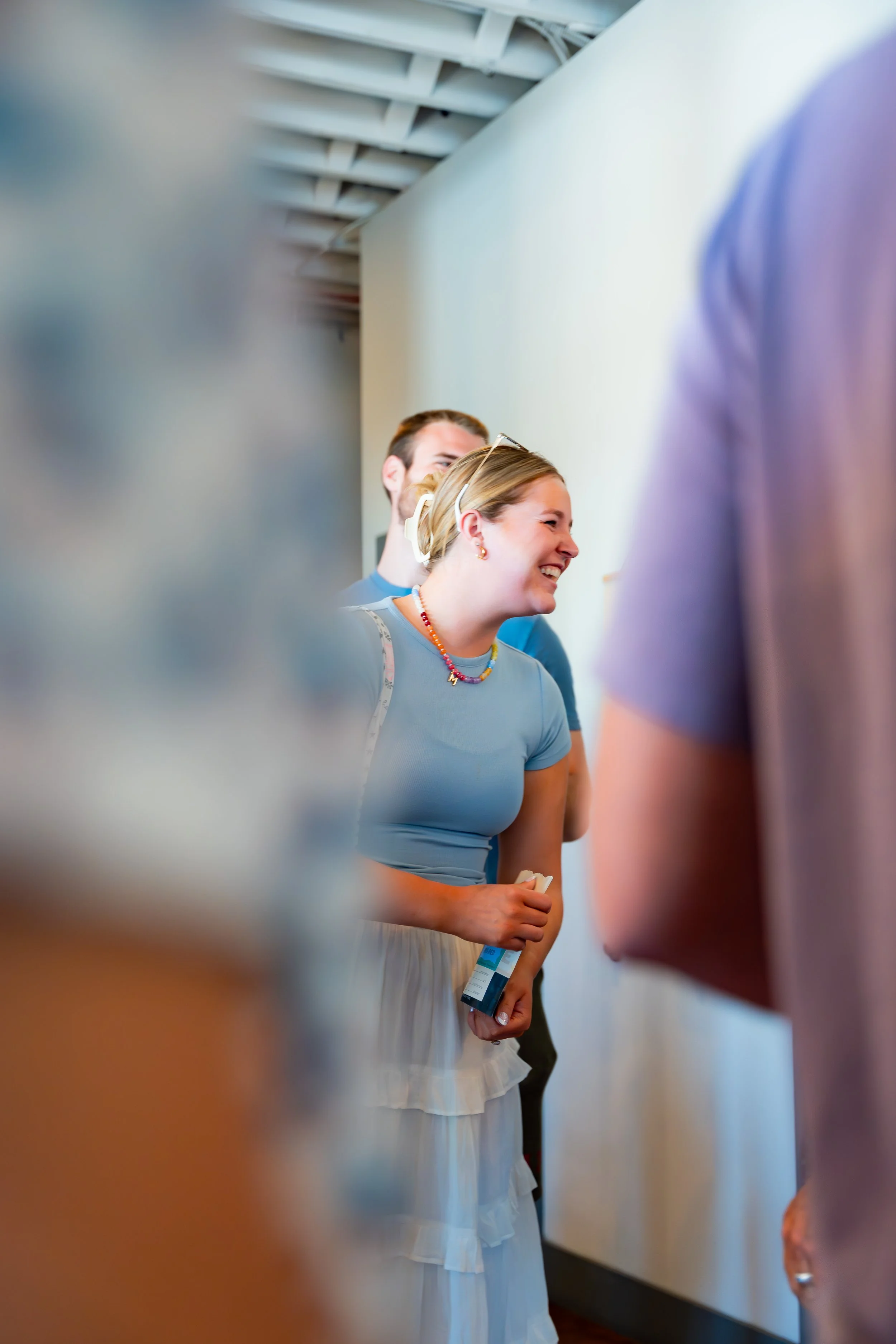 Smiling woman wearing a blue shirt, sunglasses on her head, and a colorful beaded necklace, standing among others in a brightly lit indoor space with a modern ceiling and white wall.