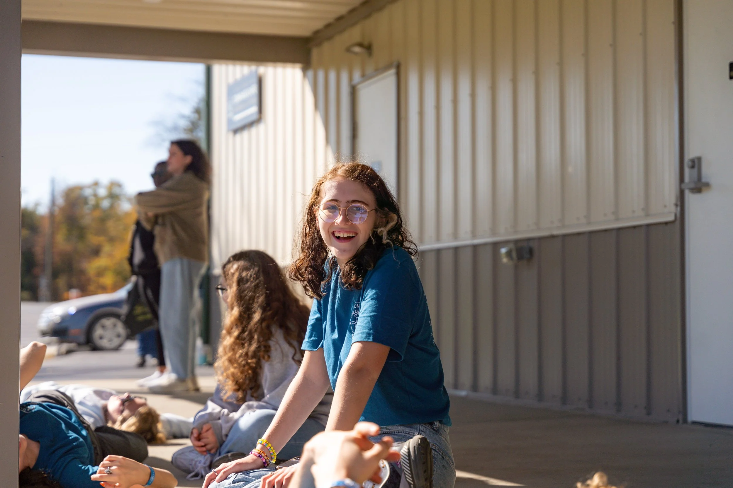 A girl with curly brown hair, glasses, and bracelets, smiling and talking to others while sitting on the ground outside a building with a metal exterior wall. Other people are in the background, some standing and some sitting, with cars parked behind