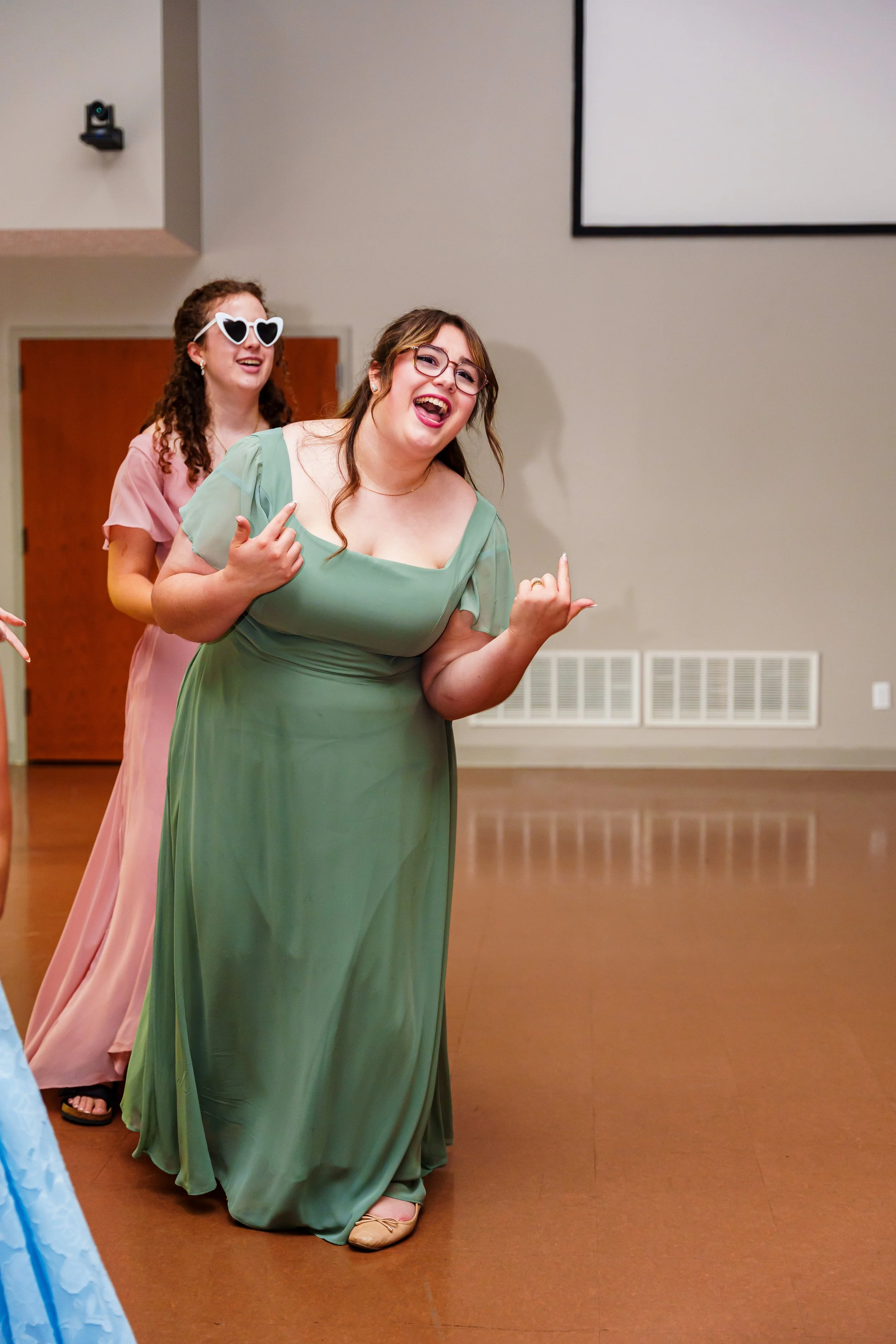 Two women in evening gowns, one in a green dress and the other in a pink dress, are enjoying a dance at a celebration or event.