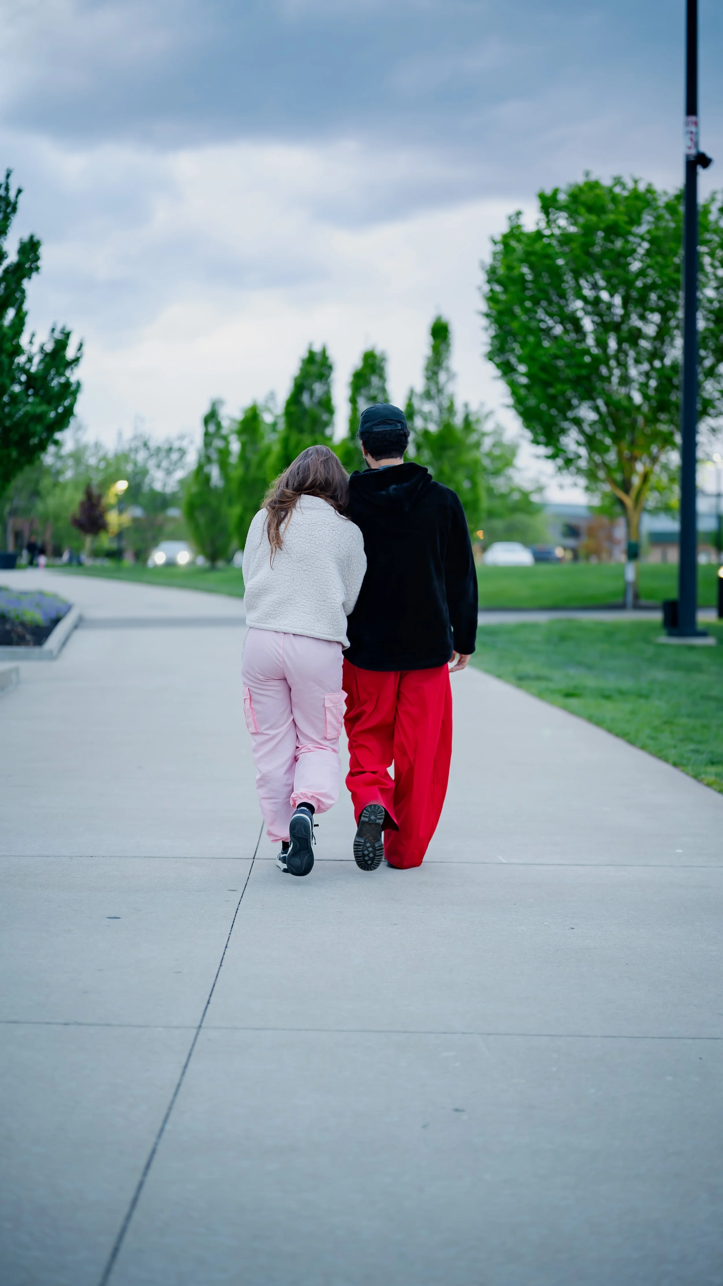 Two people, a woman and a man, walk arm in arm on a sidewalk in a park with green trees, under a cloudy sky.