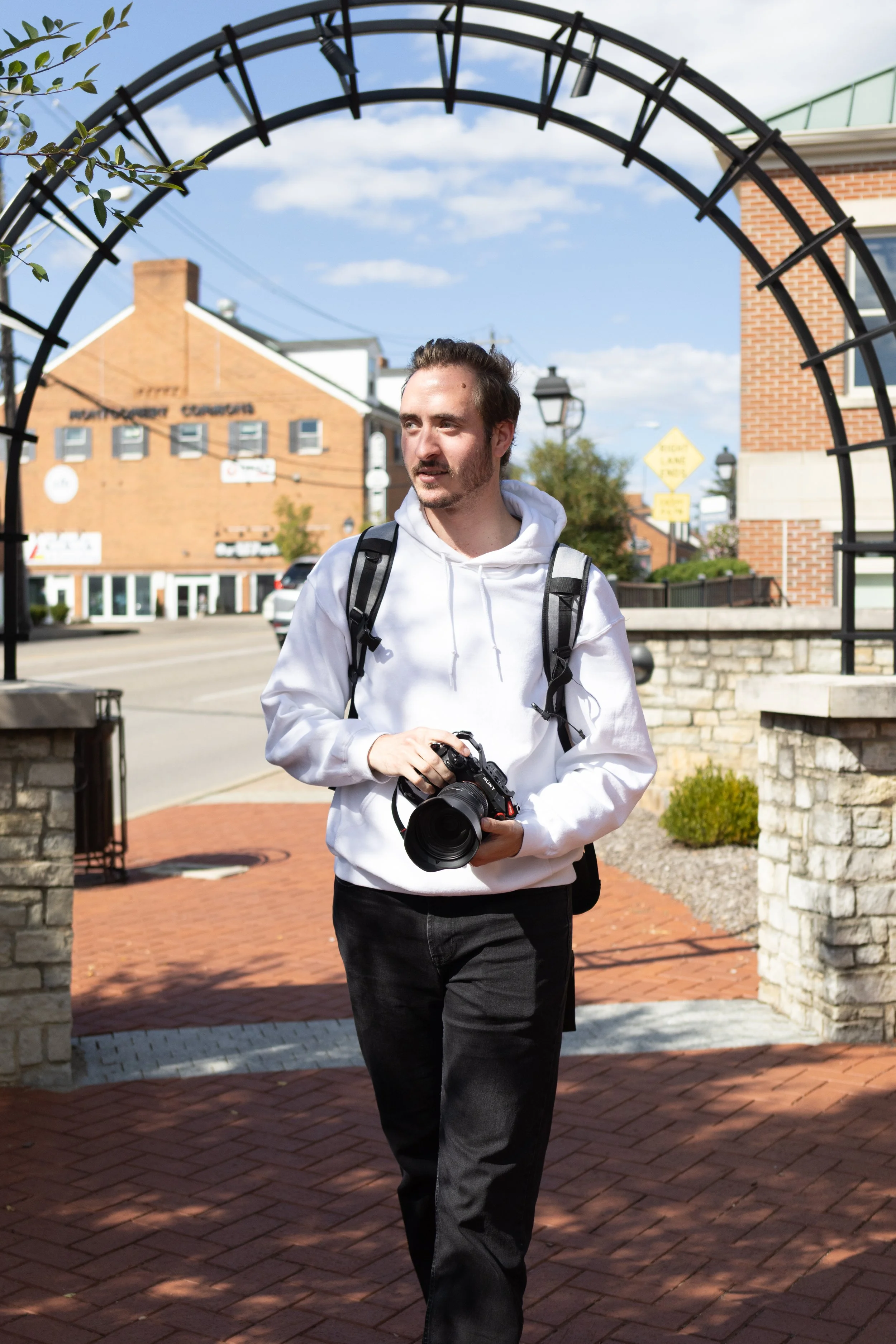 A young man with brown hair and a beard, wearing a white hoodie and black pants, holding a camera, walking outdoors through an archway on a sunny day in a small town or city with brick buildings and a blue sky.