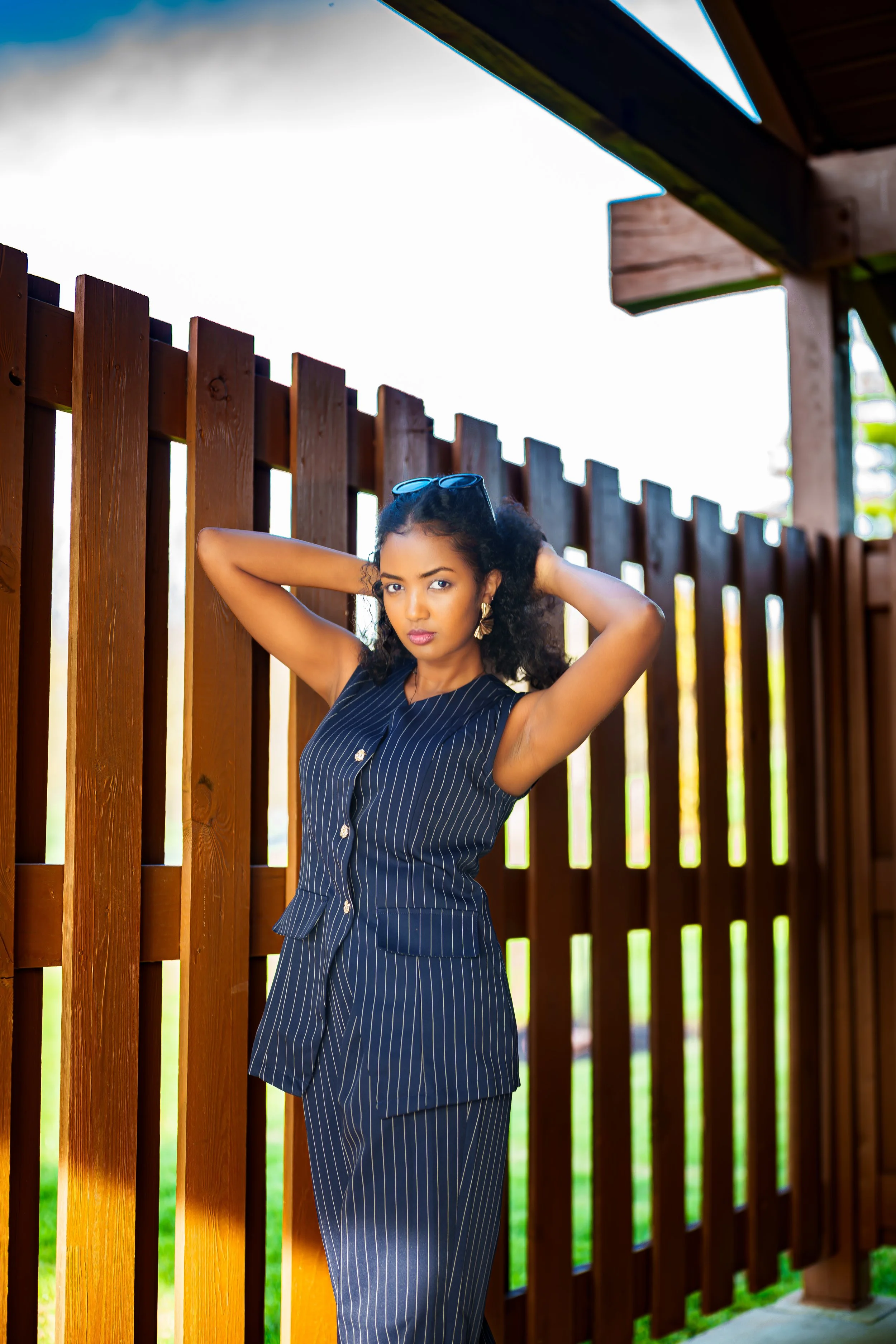 A young woman with curly hair and sunglasses on her head posing outdoors next to a wooden fence, wearing a blue pinstripe sleeveless dress.