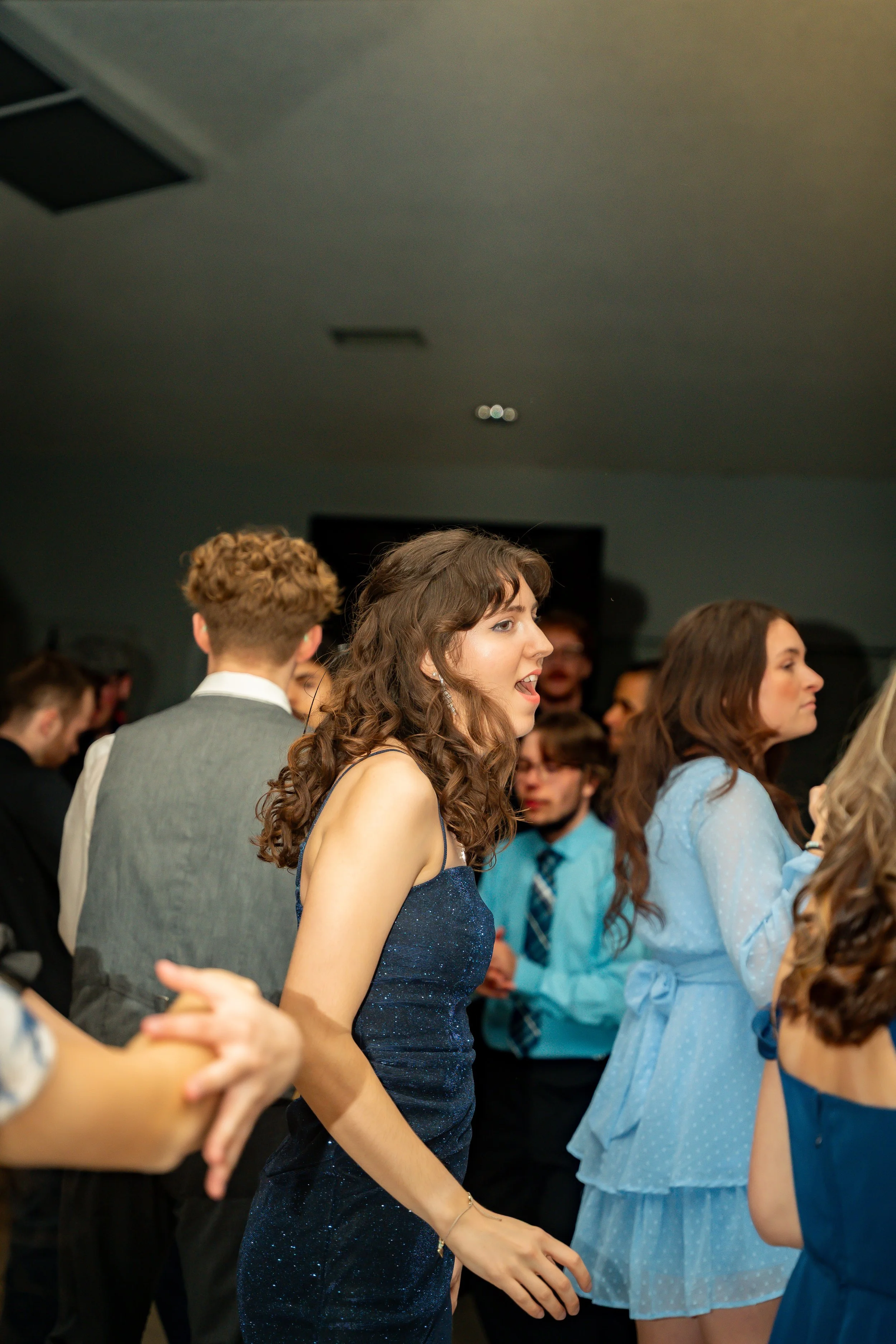 A young woman with long, curly brown hair wearing a sparkly blue dress is dancing at a social event or party with other people. She is in a line with others, holding hands or linking arms.